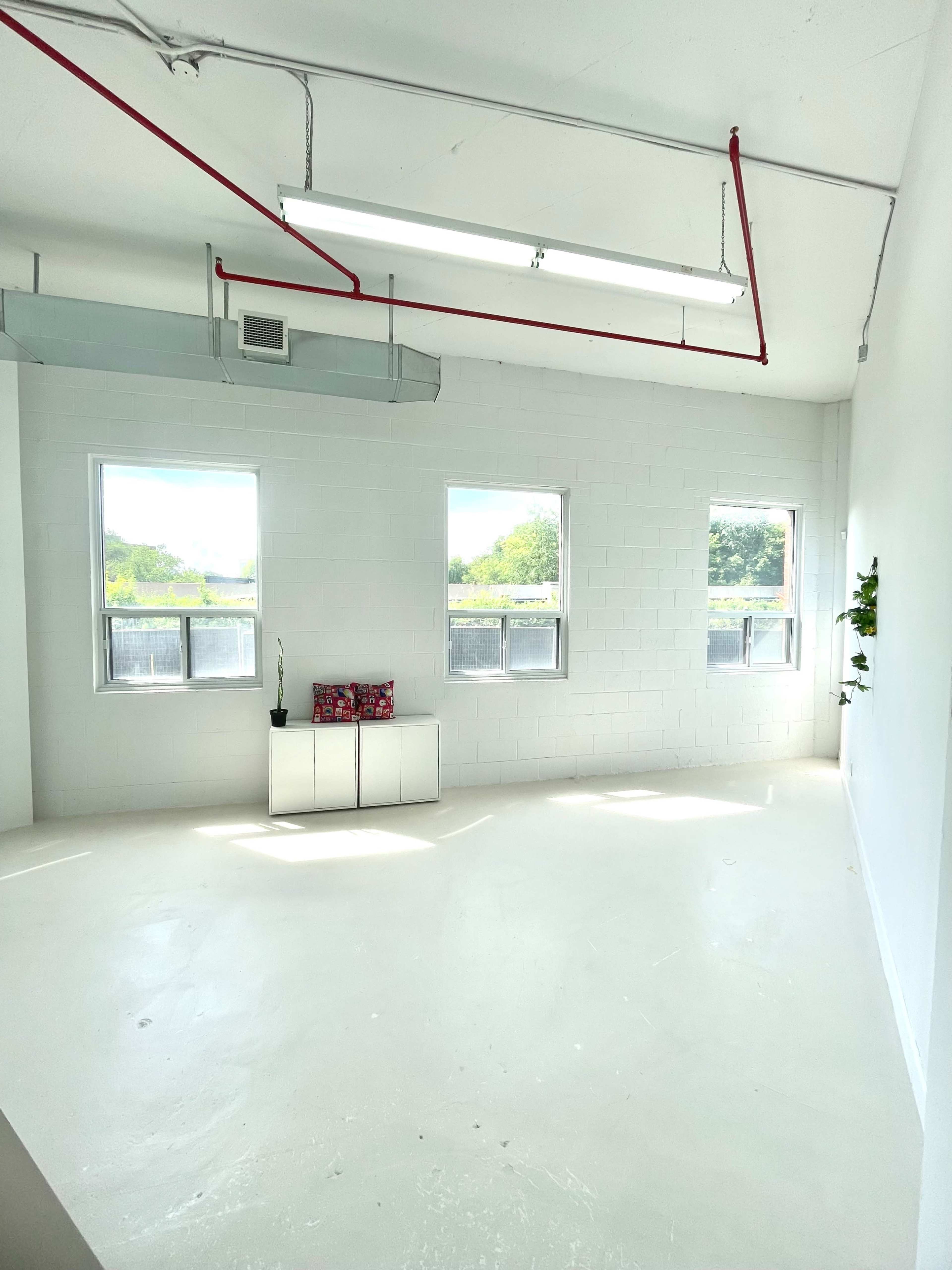 The image shows a bright, empty room with white walls, two windows, a ceiling with exposed piping, and a small table against one wall.