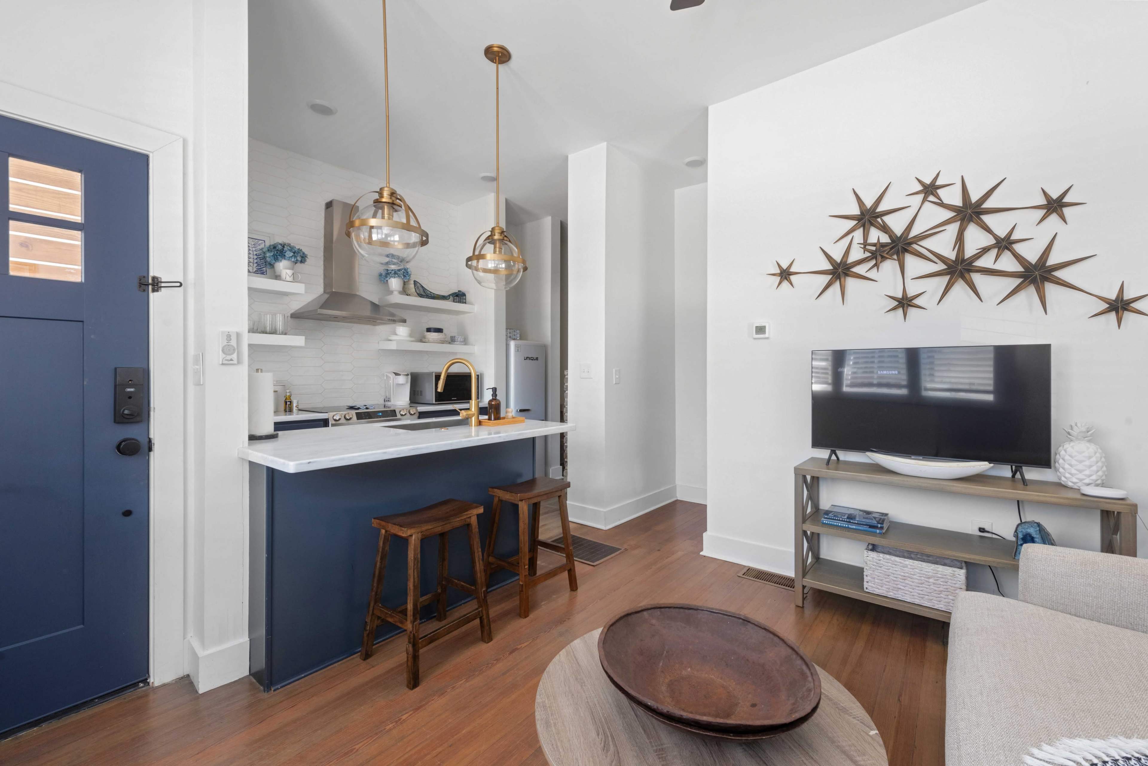 The image shows a modern, open-concept living space featuring a kitchen bar with two stools, a television on a light-colored console, and decorative star wall art.