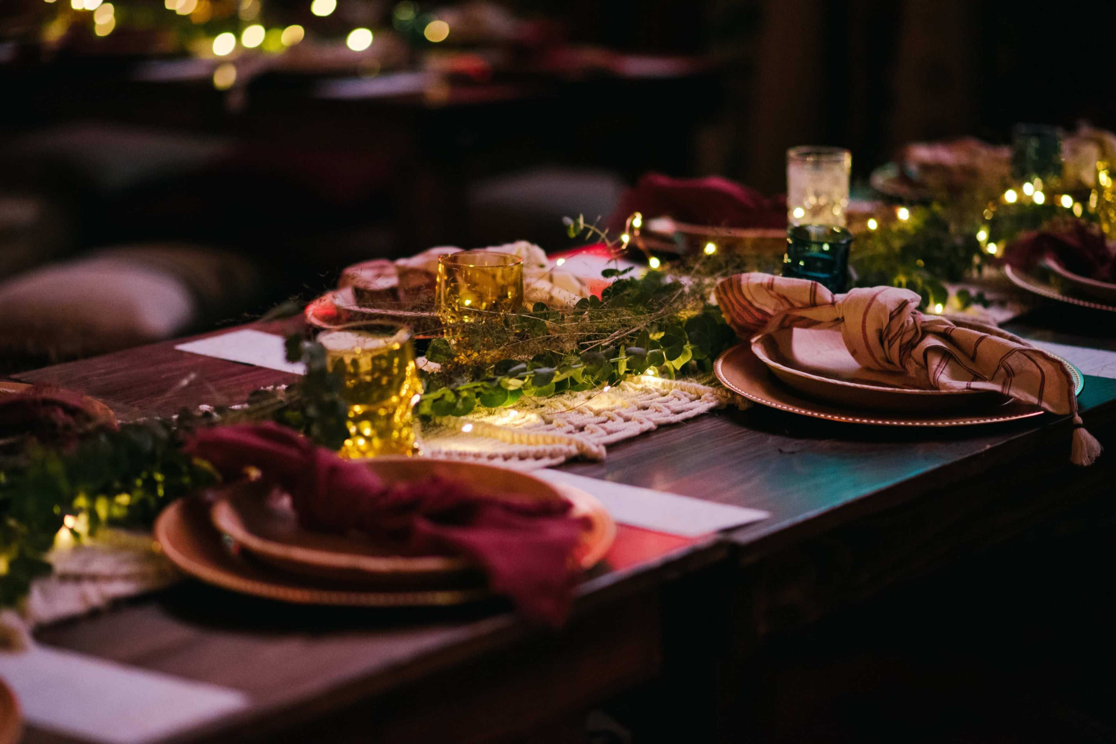 A long dining table is set with plates, candles, and green foliage, illuminated by string lights in a cozy setting.