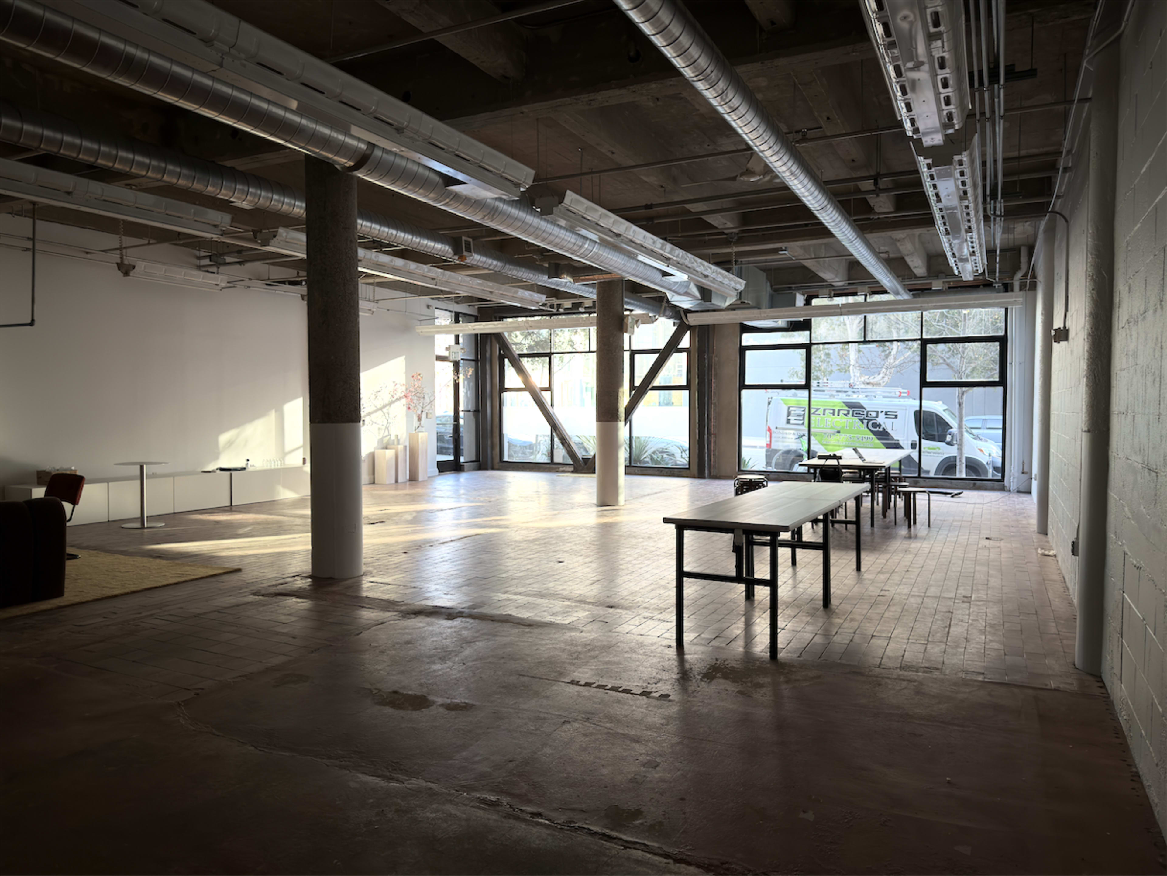 The image shows a spacious, empty room with wooden flooring, large windows, and several tables arranged in the center, with a truck visible outside.