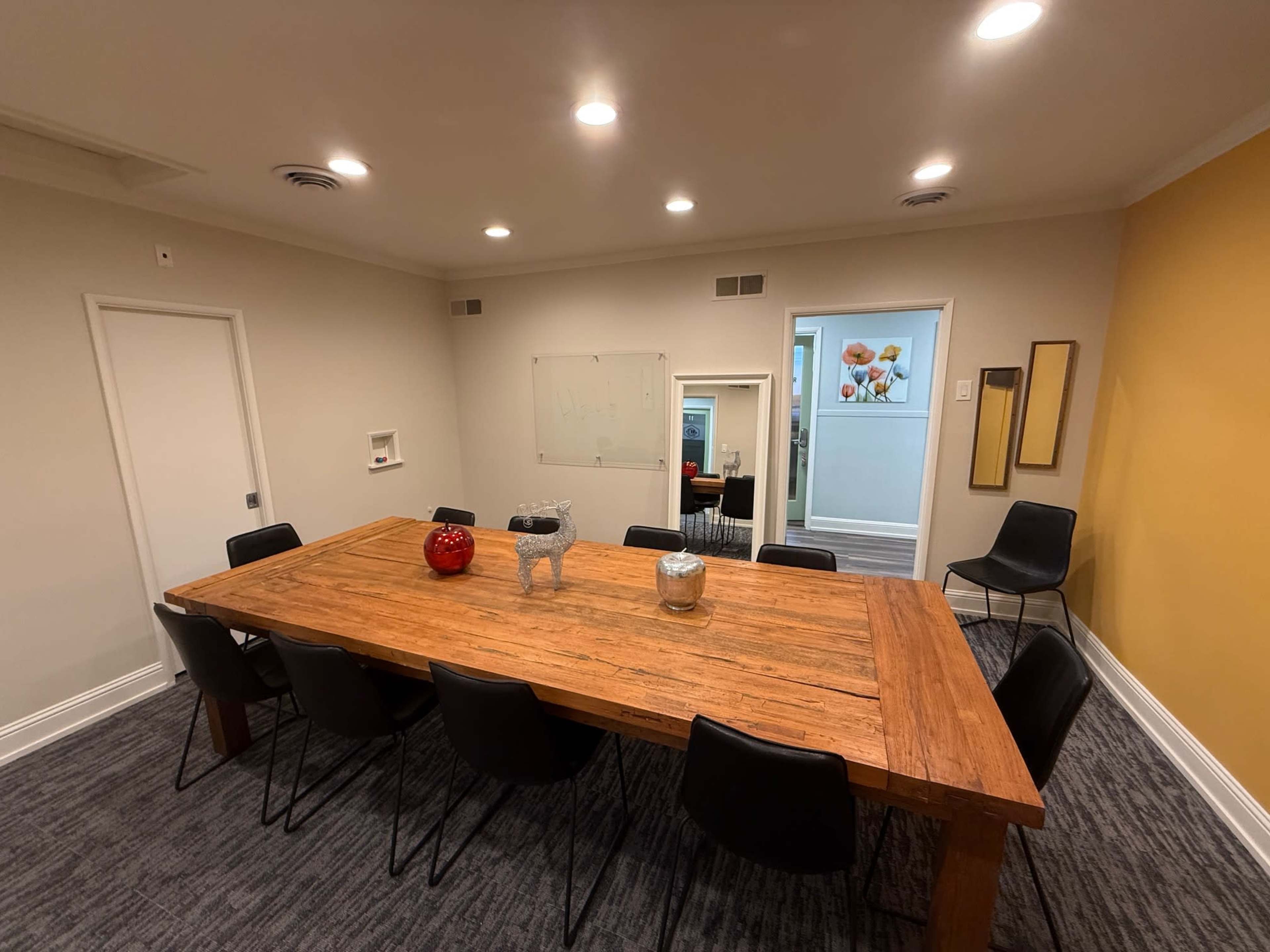 The image shows a spacious meeting room with a large wooden table surrounded by black chairs, illuminated by ceiling lights, and featuring a yellow accent wall.