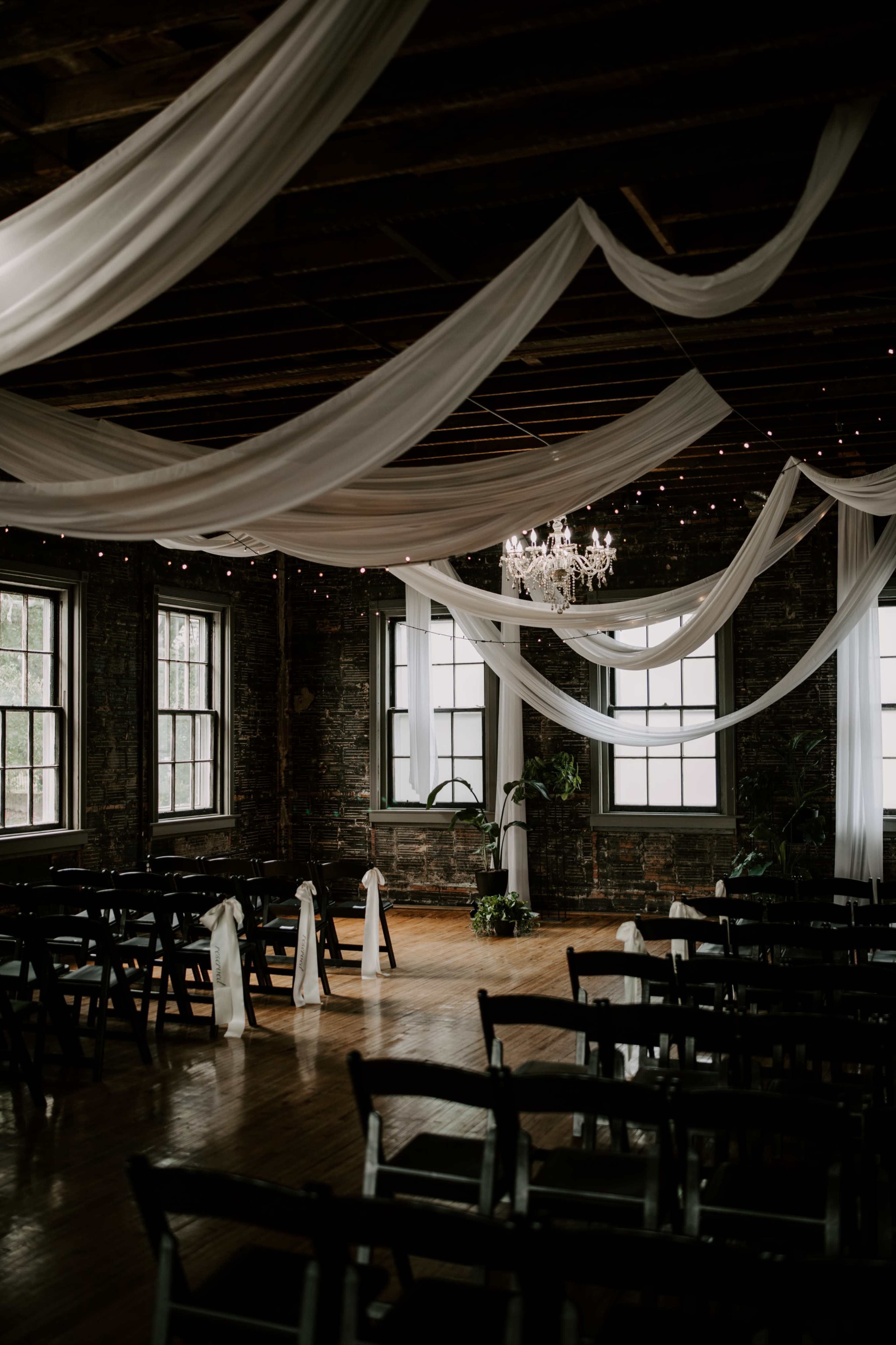 The image depicts an elegantly decorated indoor space with draped white fabric, a chandelier, and rows of chairs arranged for an event.