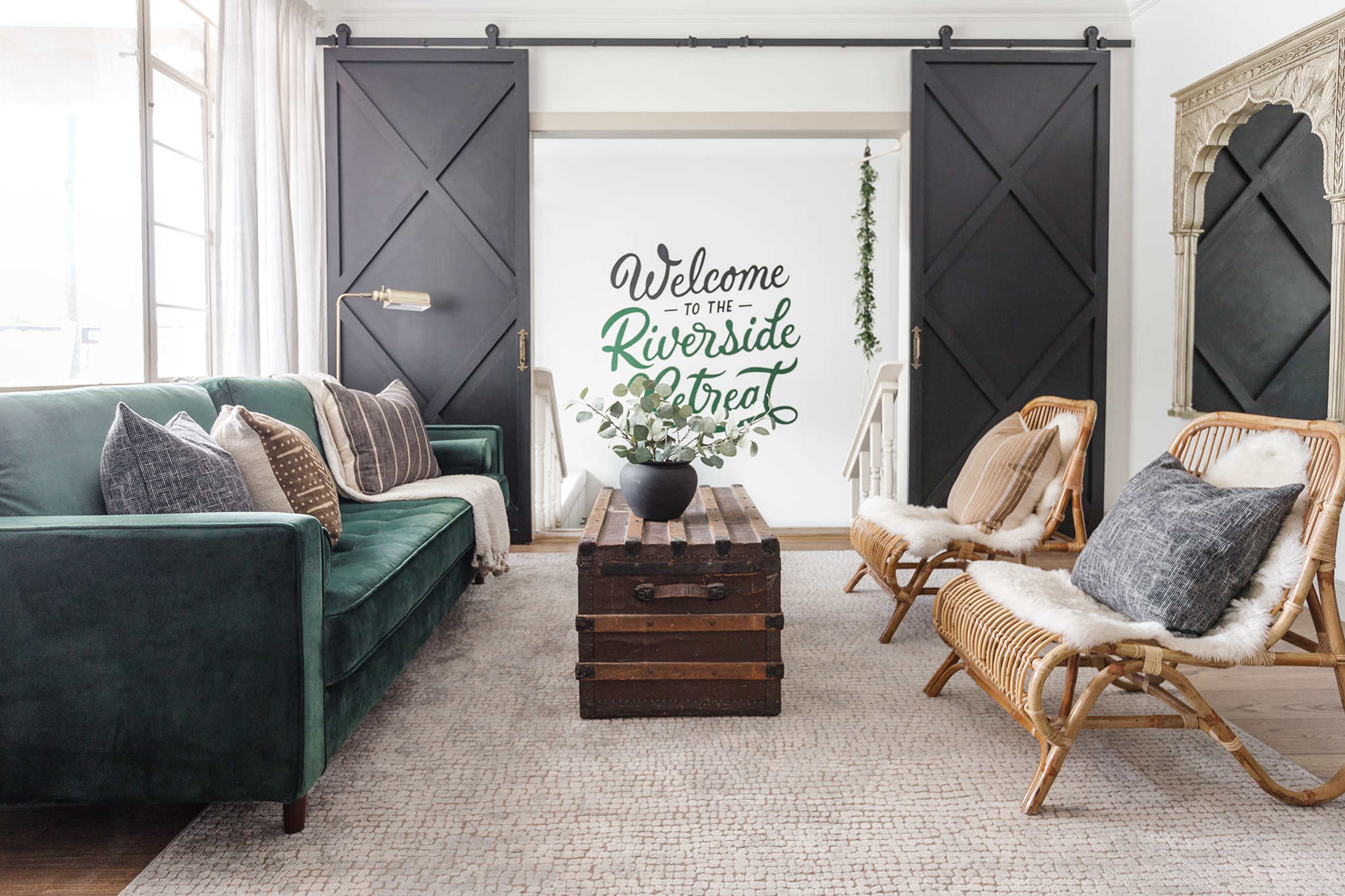A living room with a green sofa, two rattan chairs, and a wooden coffee table, featuring a wall with the words "Welcome to the Riverside Street" painted on it.