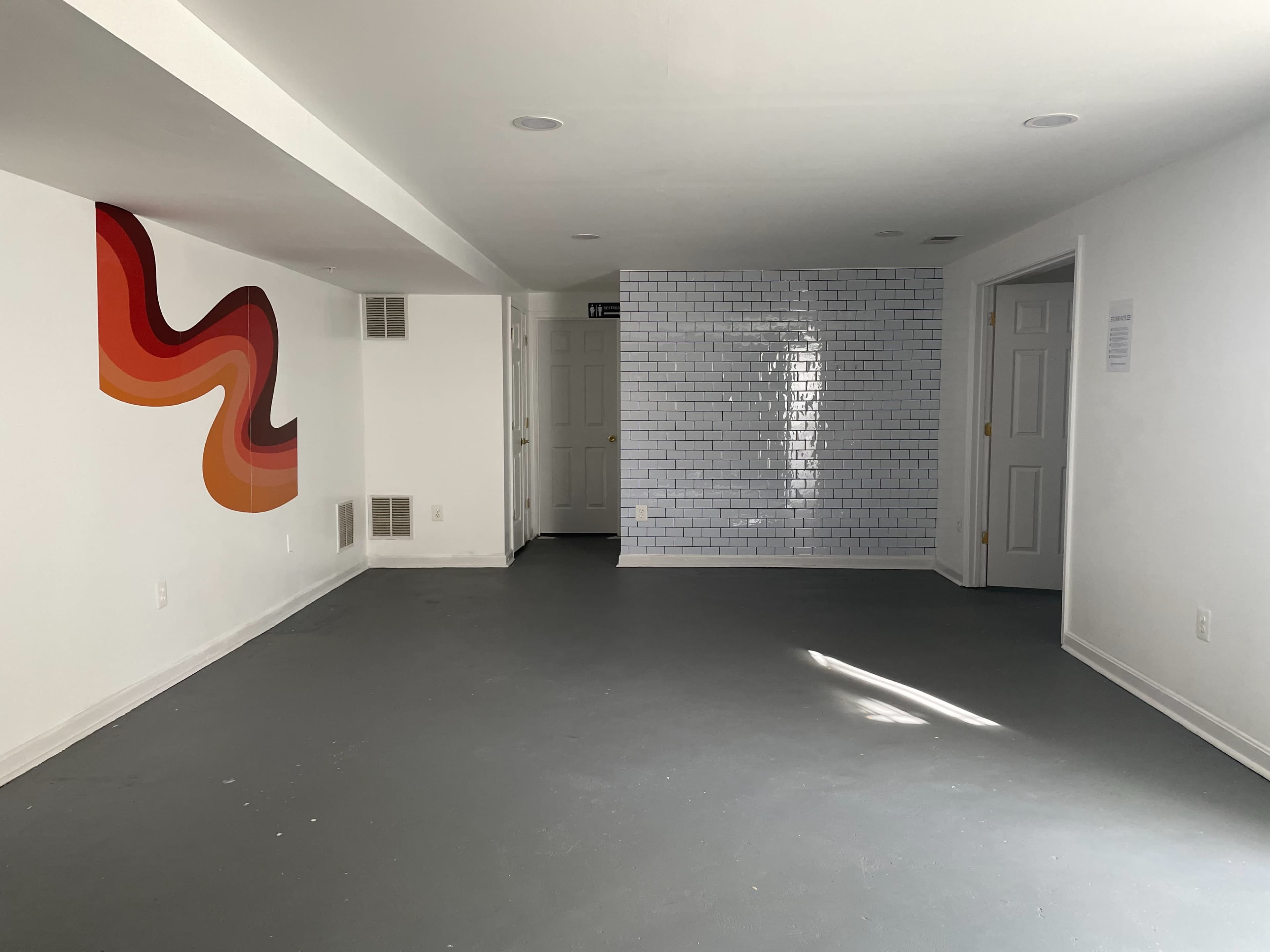 A large, empty room with a colorful abstract mural on one wall, a tiled section on another, and white walls and gray flooring.