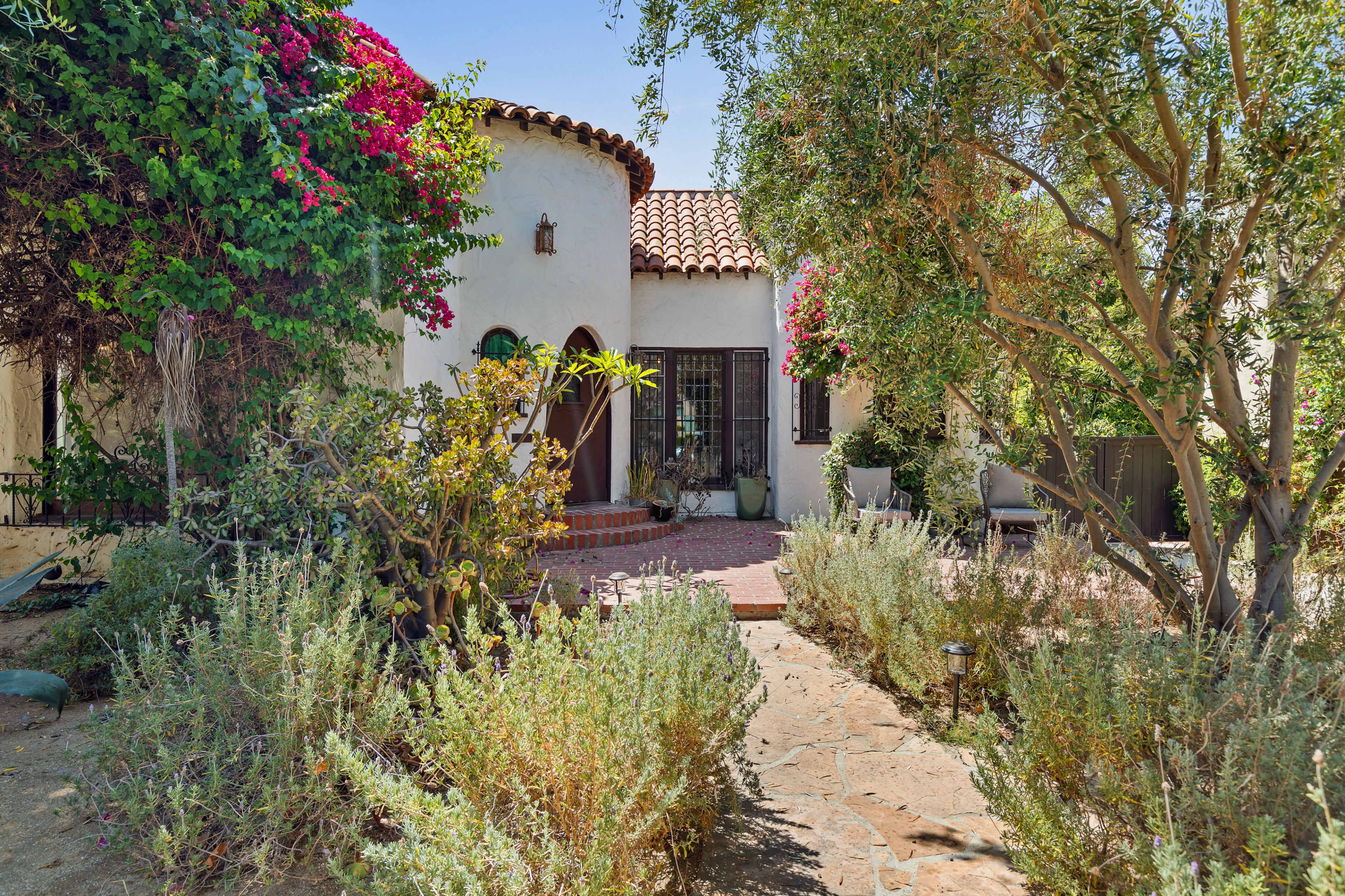 A Spanish-style house with a tiled roof is surrounded by blooming plants and a stone pathway.