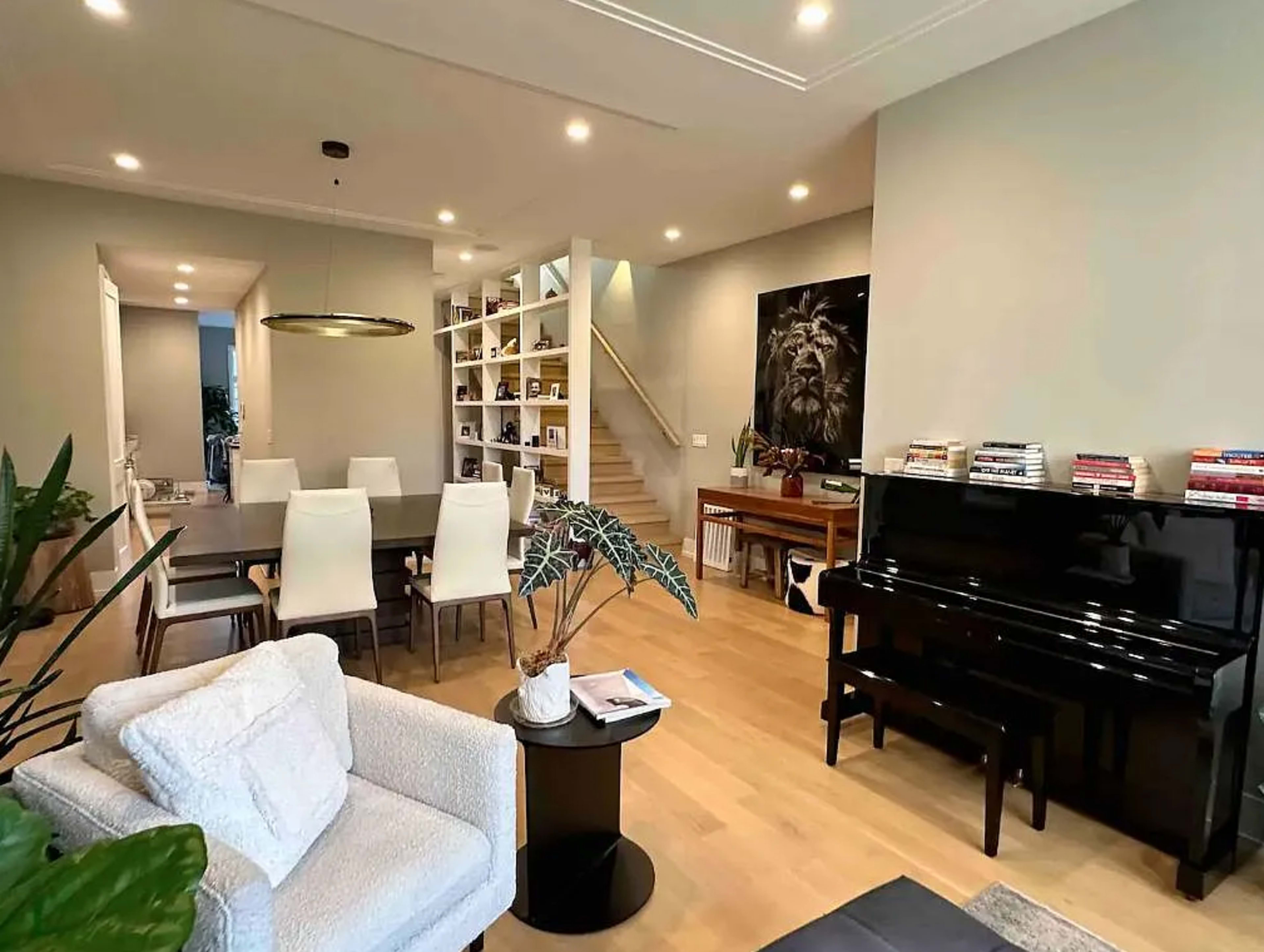 The image shows a modern living space featuring a dining area with a long table, a piano in the corner, bookshelves on one wall, and a staircase leading to an upper level.