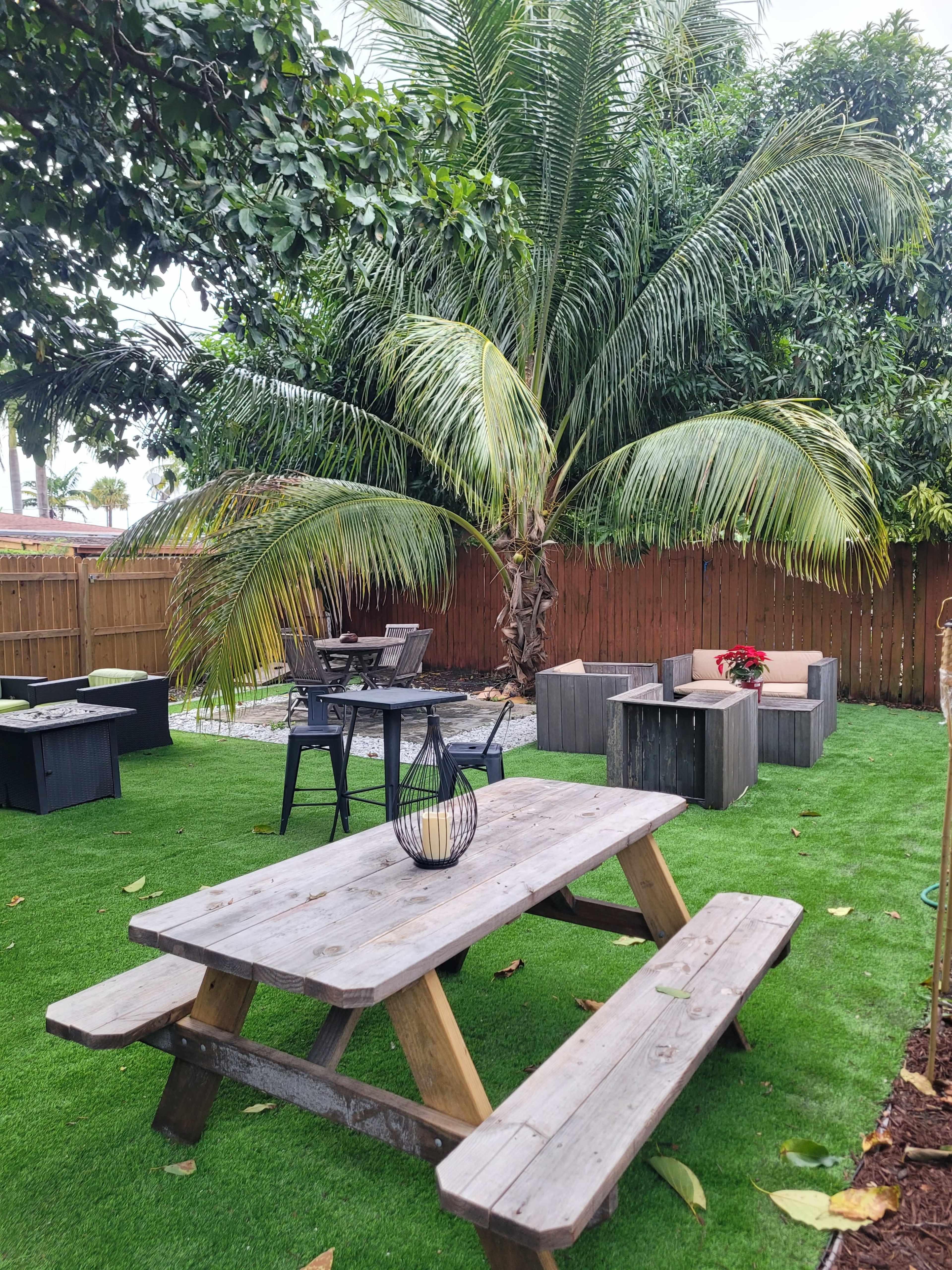 A wooden picnic table sits on a grassy area surrounded by palm trees and outdoor seating arrangements.