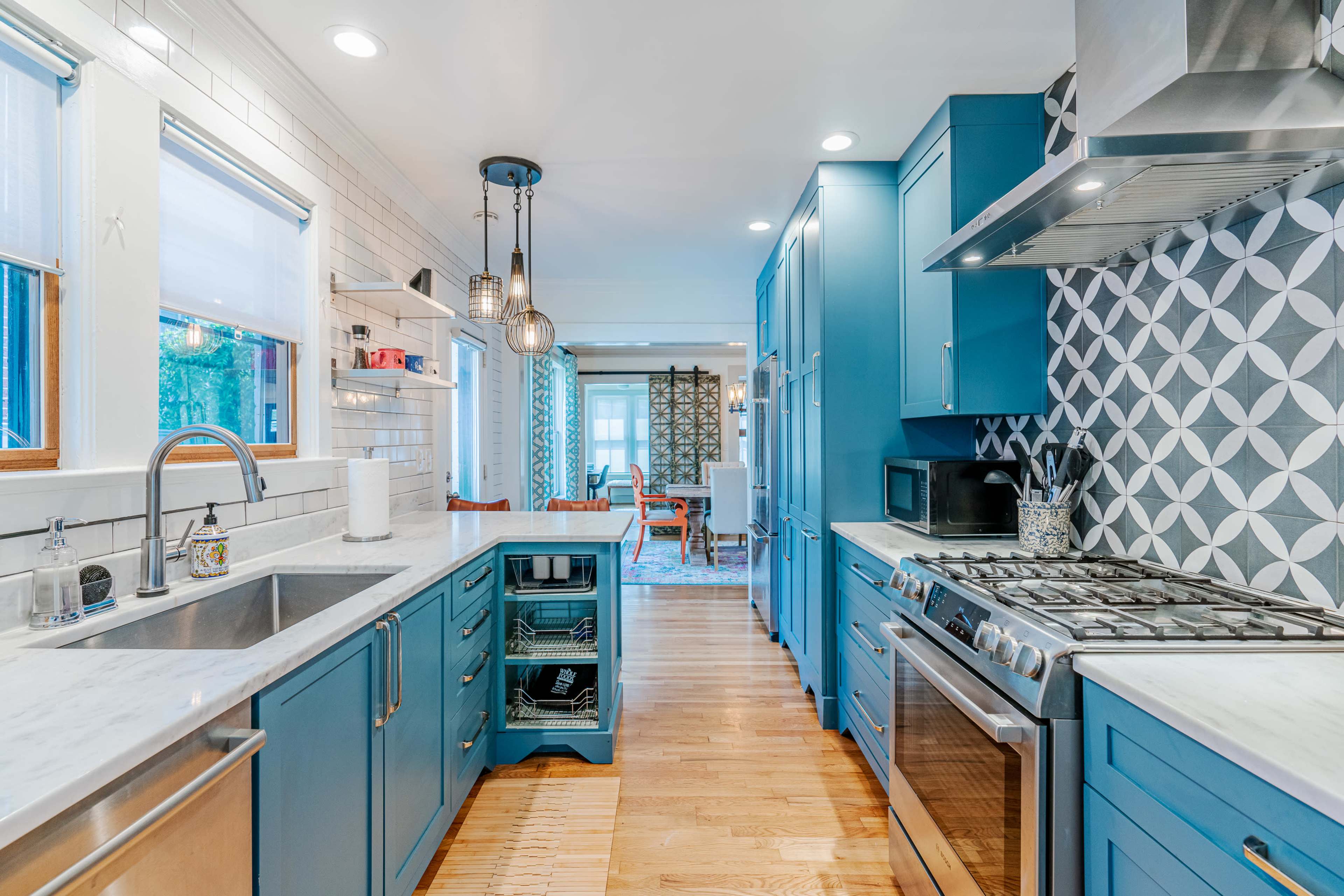 A modern kitchen with blue cabinets, a marble countertop, and stainless steel appliances, leading into a dining area with patterned curtains.