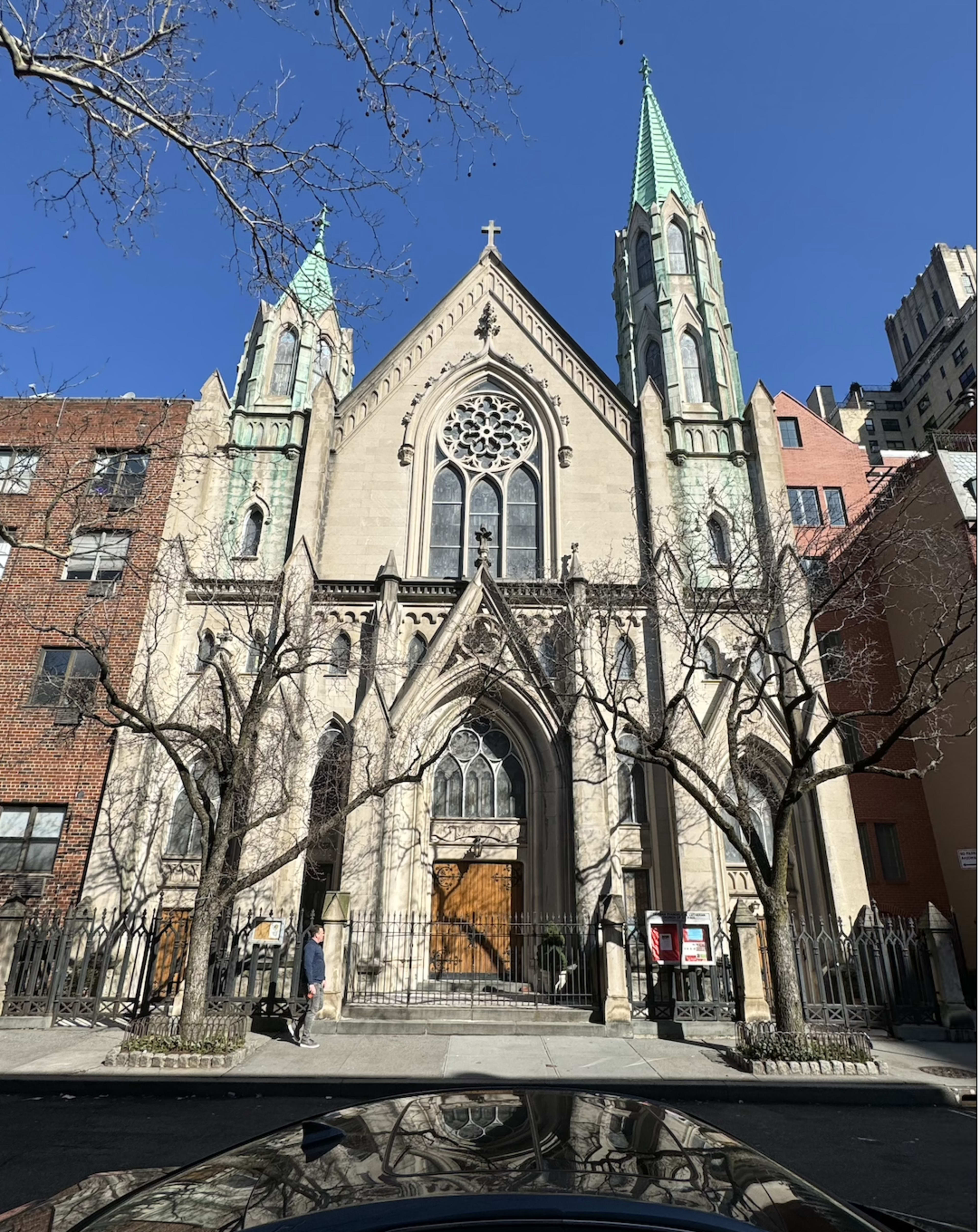 A Gothic-style church with pointed towers and intricate stained glass windows, located between urban buildings against a clear blue sky.
