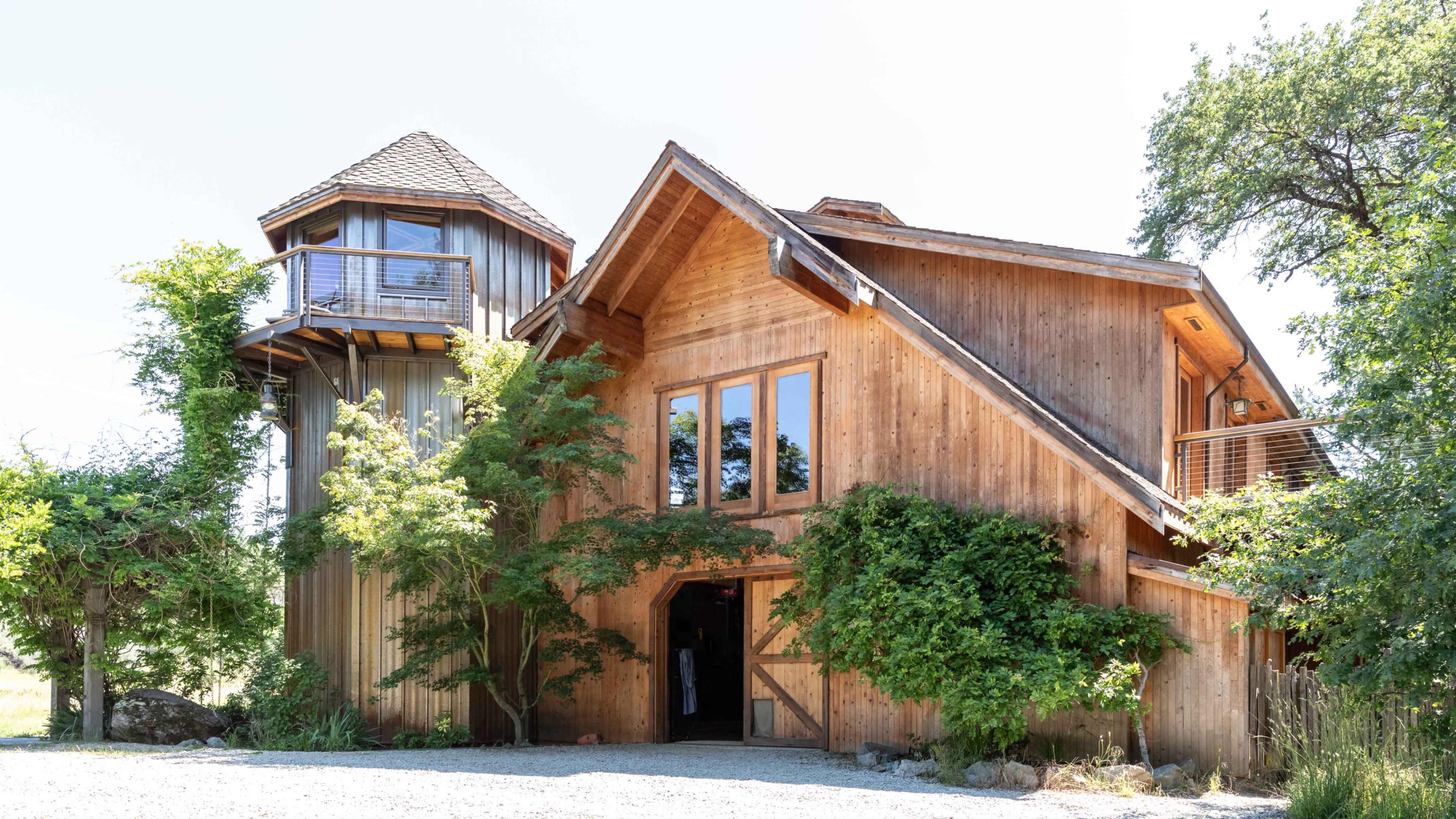 A wooden house with a multi-level design, featuring large windows and a tower, is surrounded by greenery.