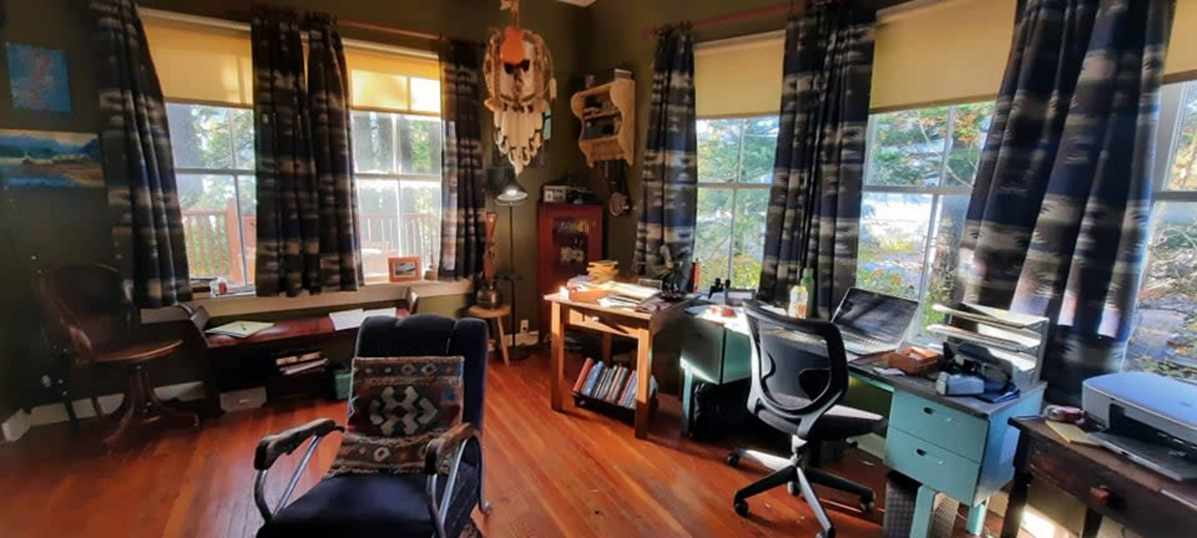 A cozy home office features a mix of wooden furniture, large windows with light filtering through, and a variety of books and papers scattered across the workspace.
