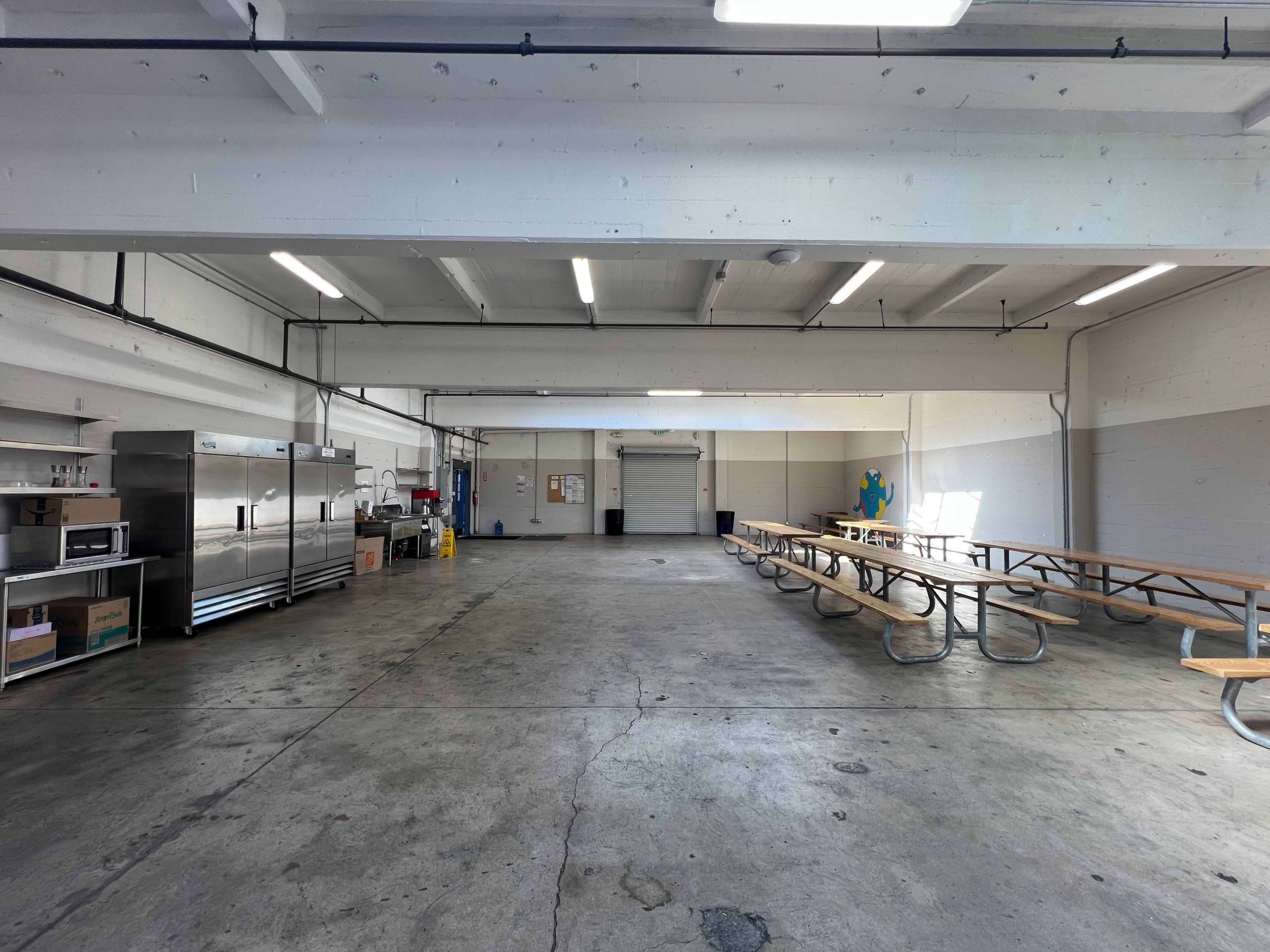 A spacious industrial kitchen area with stainless steel appliances and a long wooden picnic-style table.