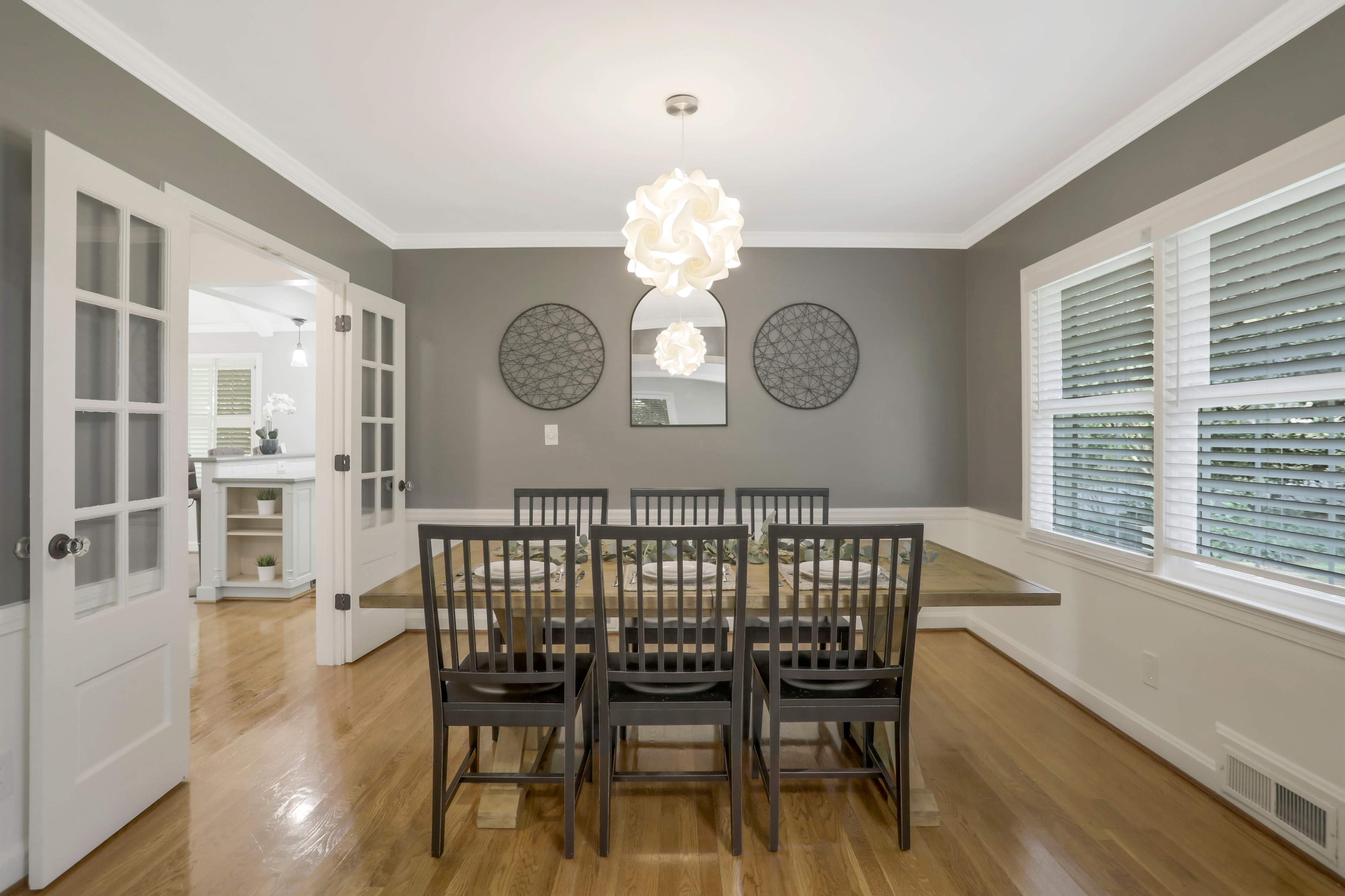 A dining room features a wooden table surrounded by six black chairs, with a modern light fixture overhead and decorative mirrors on the gray walls.