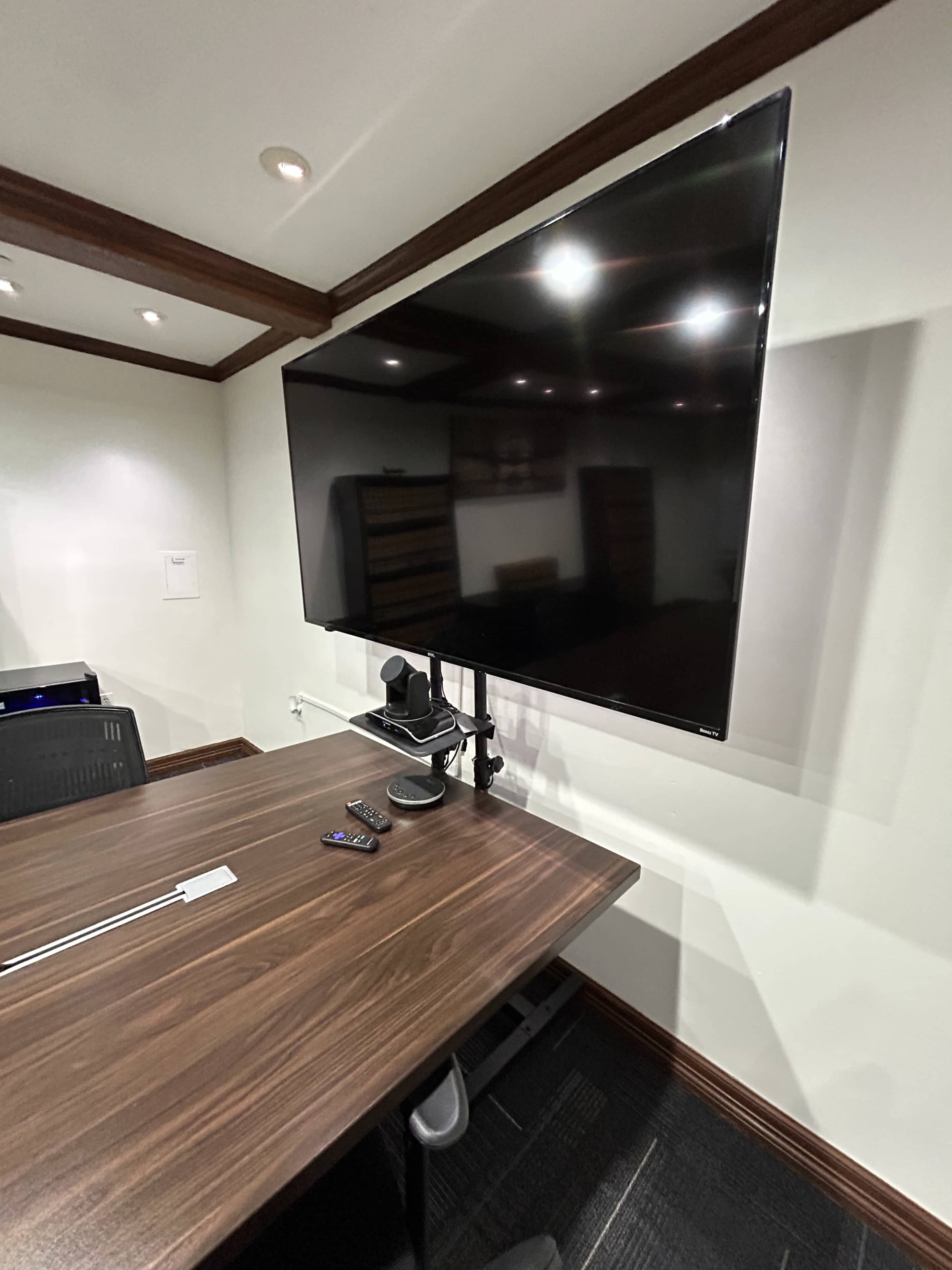 The image shows a conference room with a large flat-screen TV mounted on the wall above a wooden conference table and a video conferencing camera positioned in front of it.