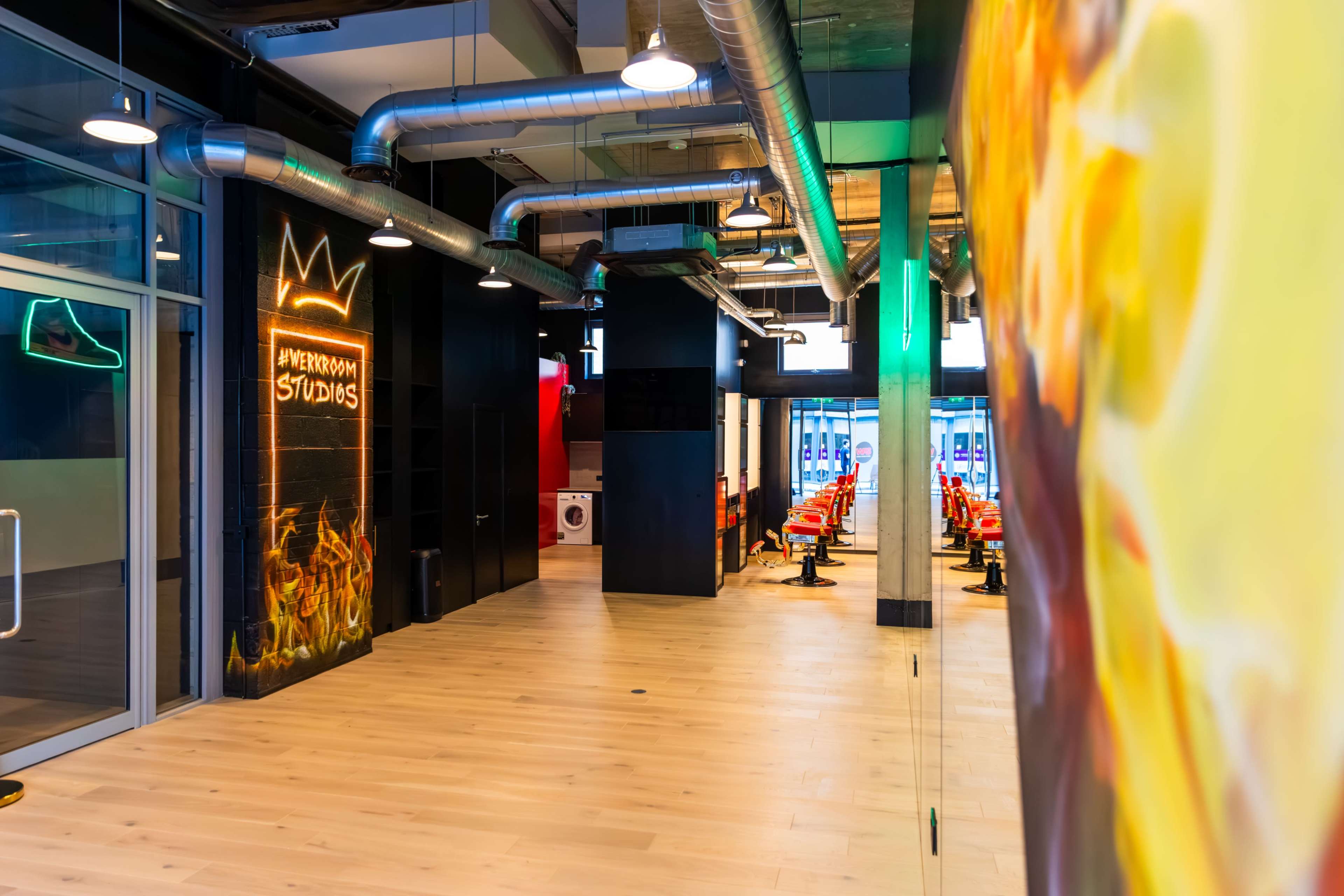 The image shows a spacious interior of a modern studio with exposed ventilation, bright colors, and a row of red chairs.
