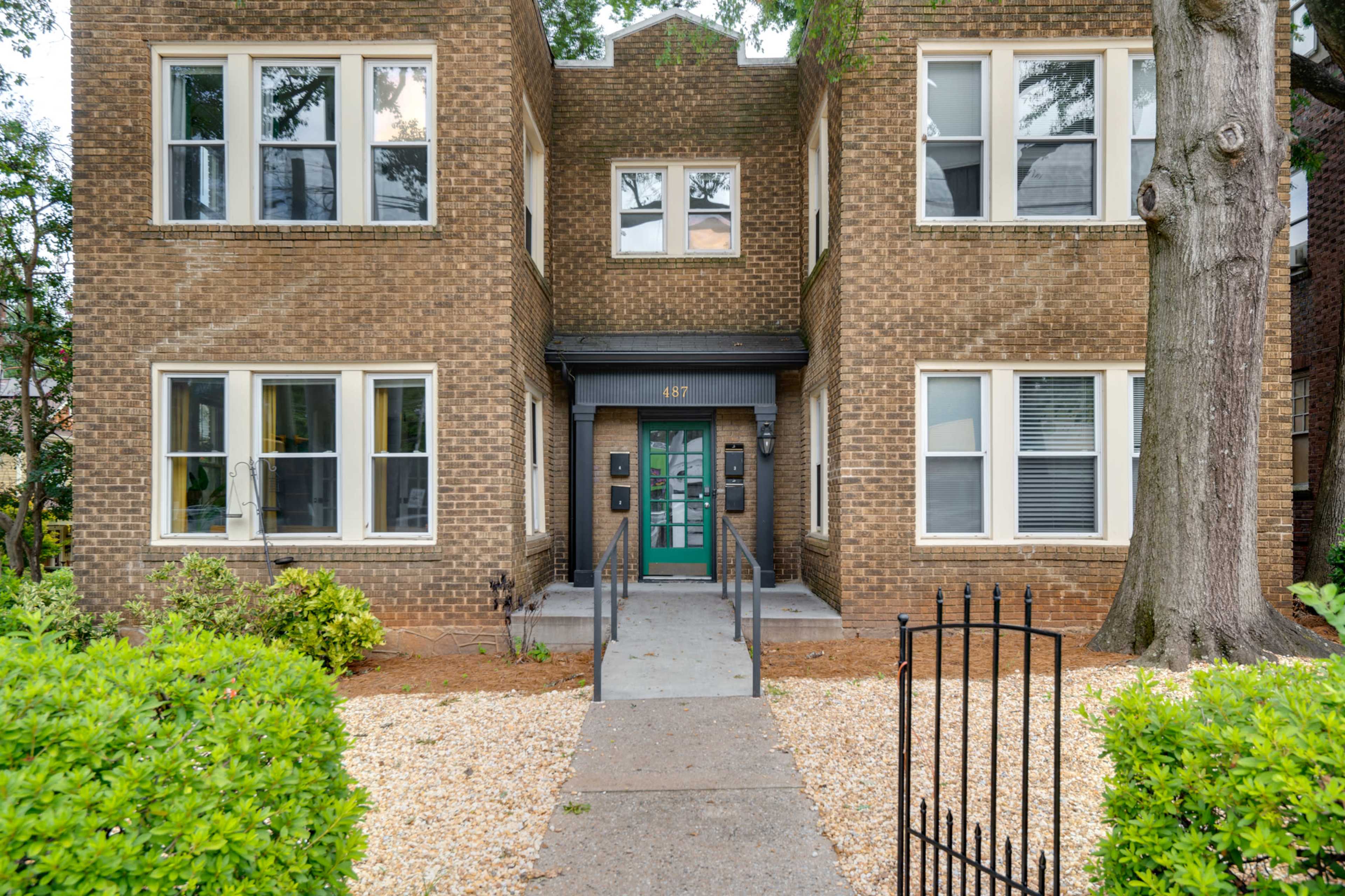 The image shows a two-story brick apartment building with a central entrance, flanked by windows and surrounded by greenery and a walkway.