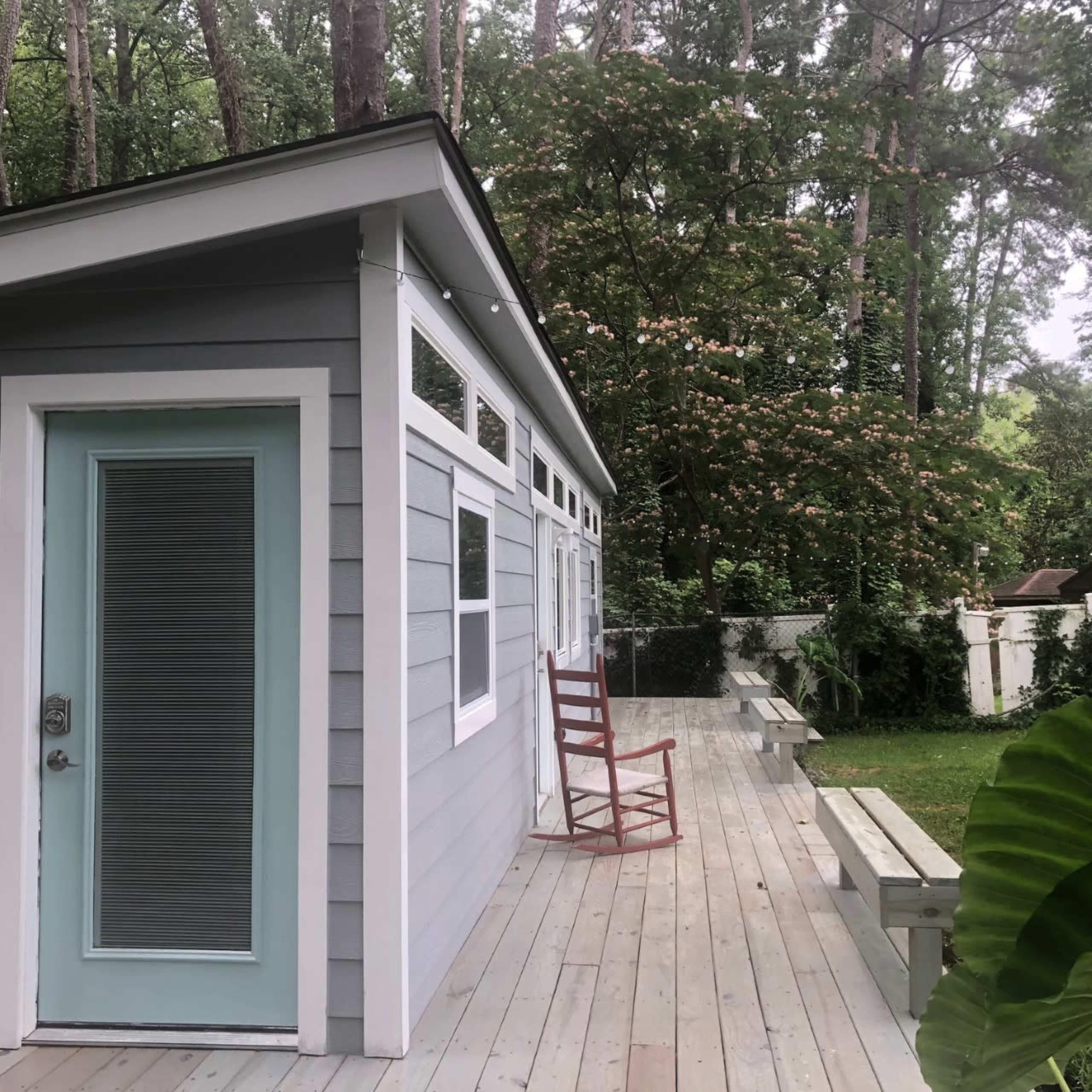 A small, modern house with a light blue door is situated on a wooden deck surrounded by trees.