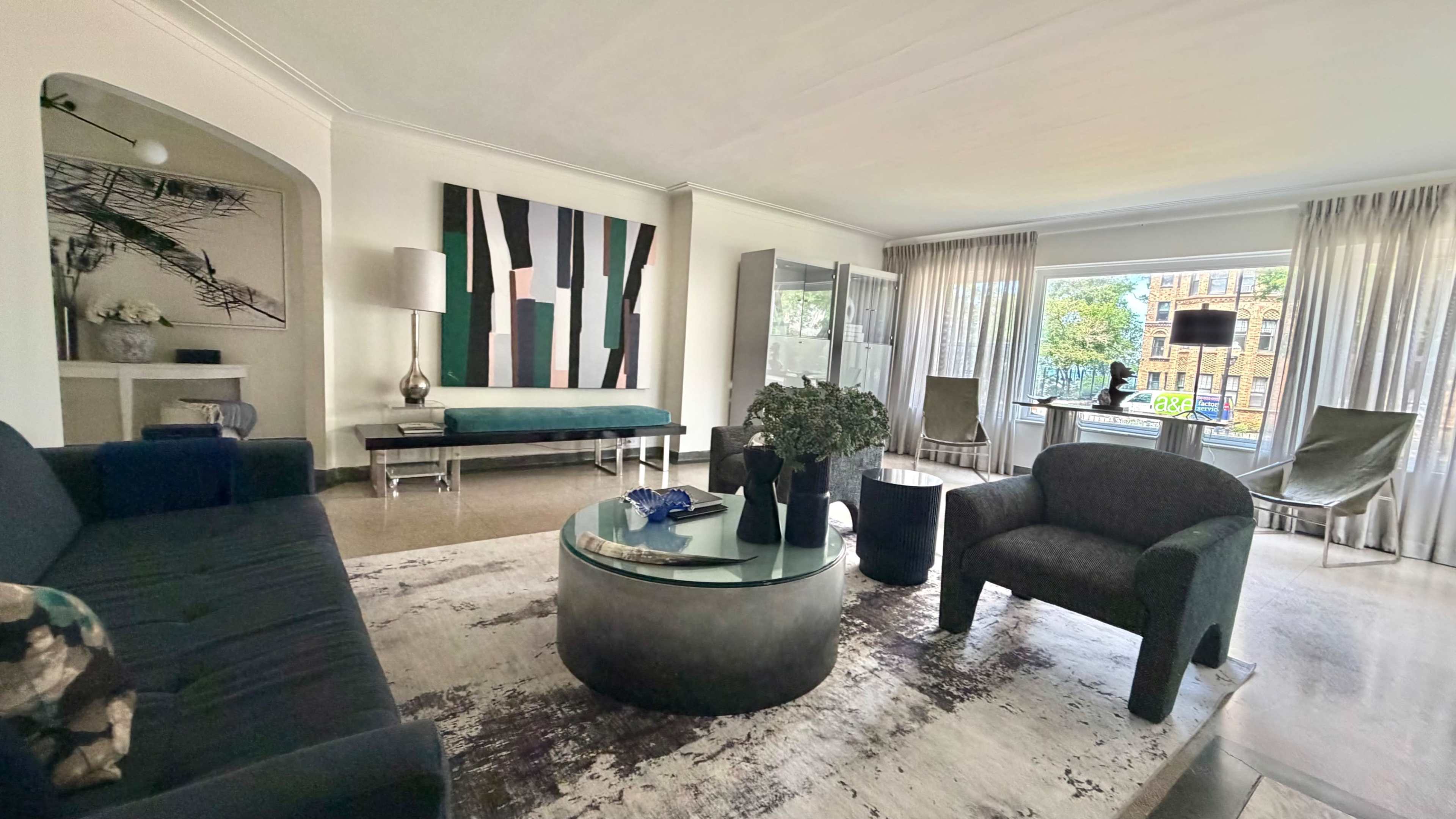 The image depicts a modern living room featuring a round glass coffee table, a large abstract painting on the wall, and a mix of seating options, including a dark sofa and armchairs, with large windows letting in natural light.