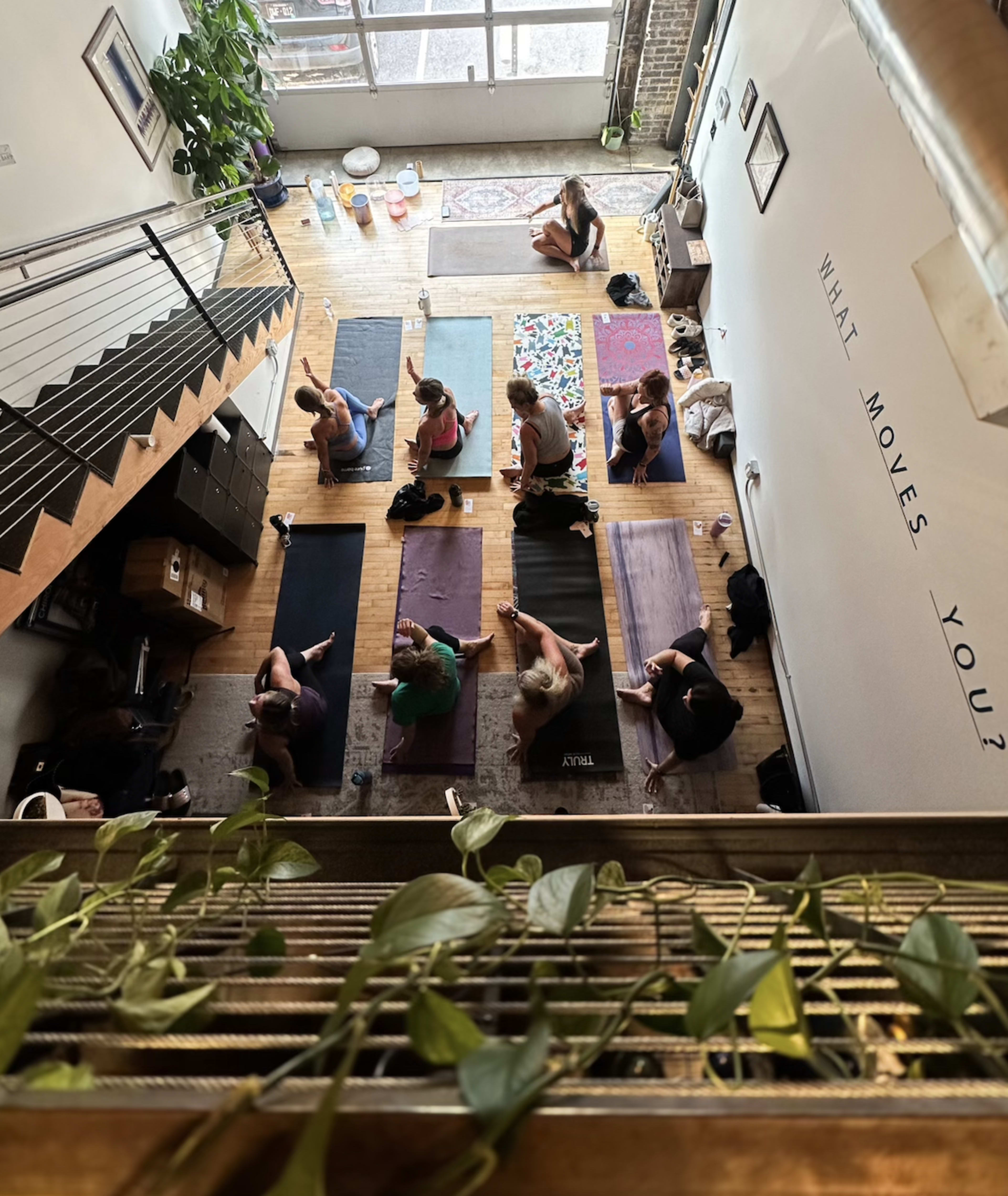 A group of people practices yoga in a studio with wooden floors and large windows.