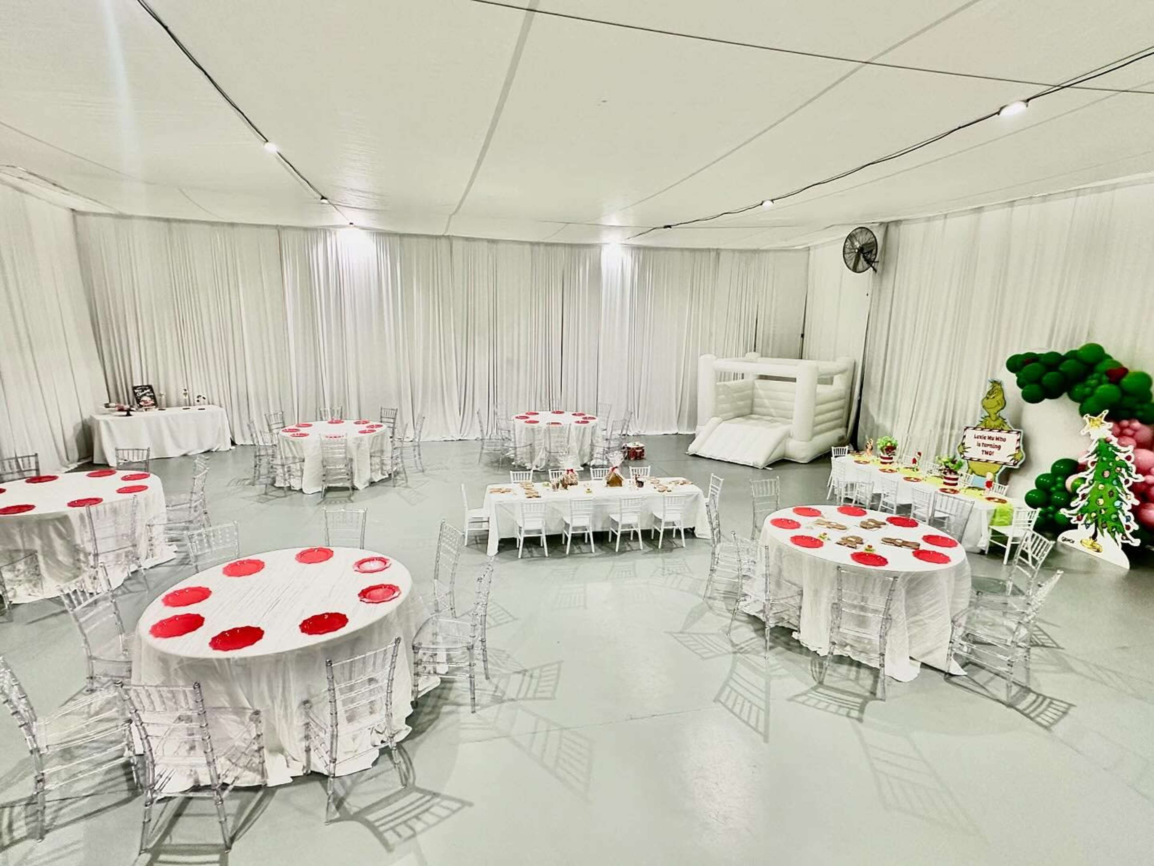 The image shows an indoor event space decorated with white drapes, round tables covered with white tablecloths and red centerpieces, and a bounce house in the corner.