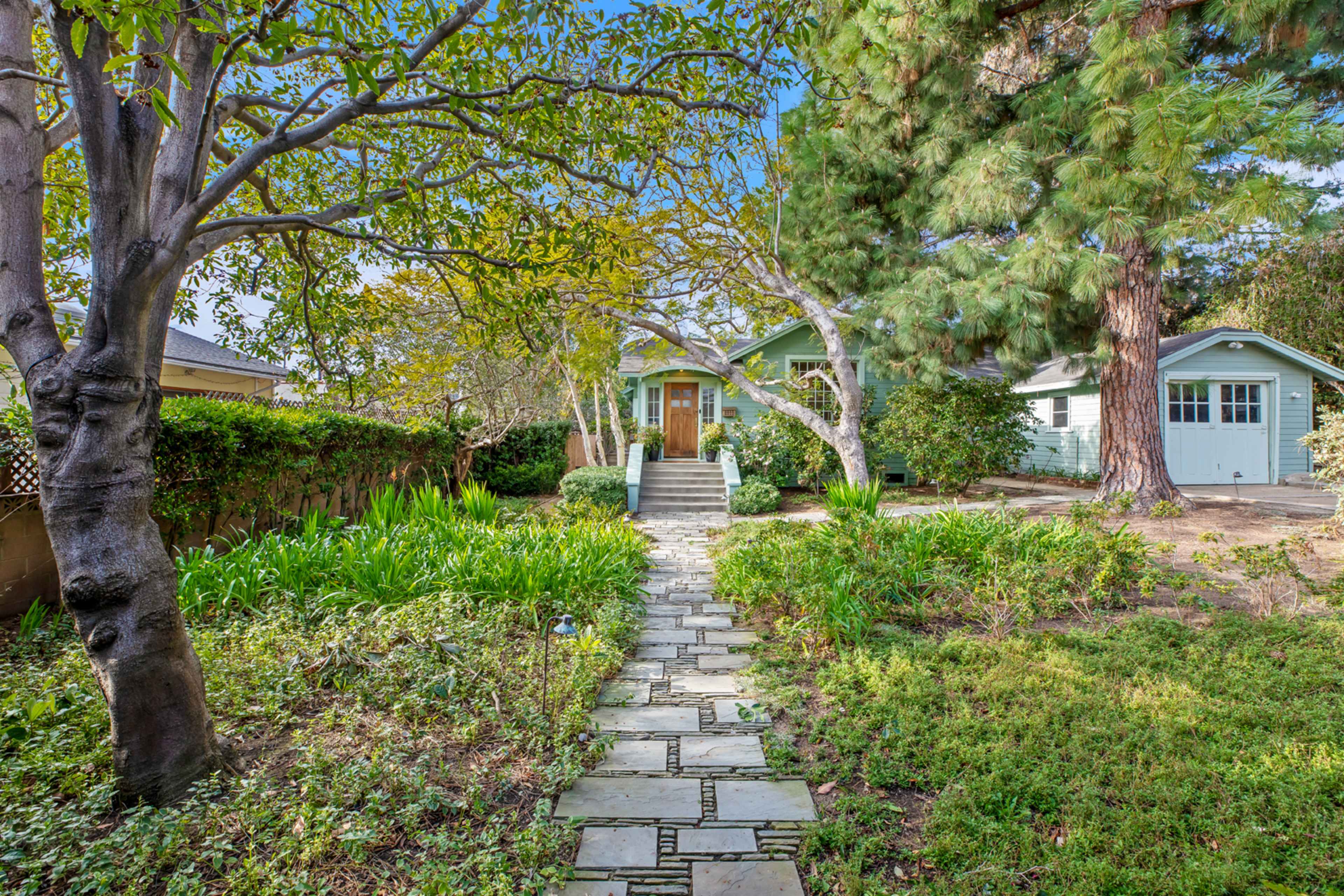 A stone pathway leads to a house surrounded by greenery and trees, with a garage visible to the right.