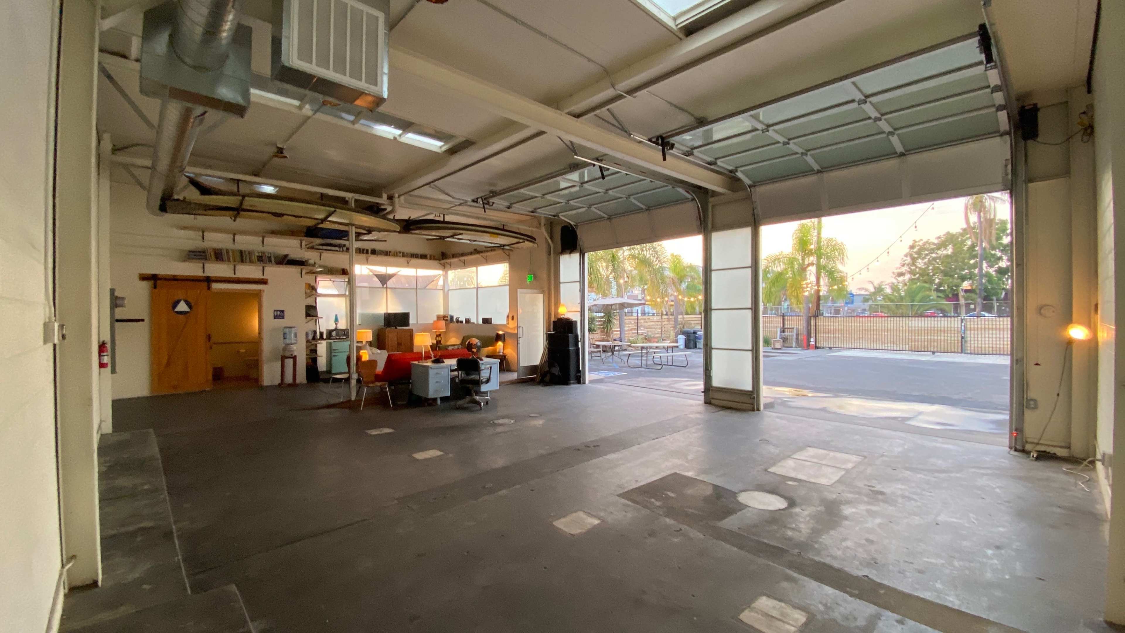 An open industrial space features a seating area with orange chairs, large garage doors, and a view of an outdoor area with a fence and greenery.