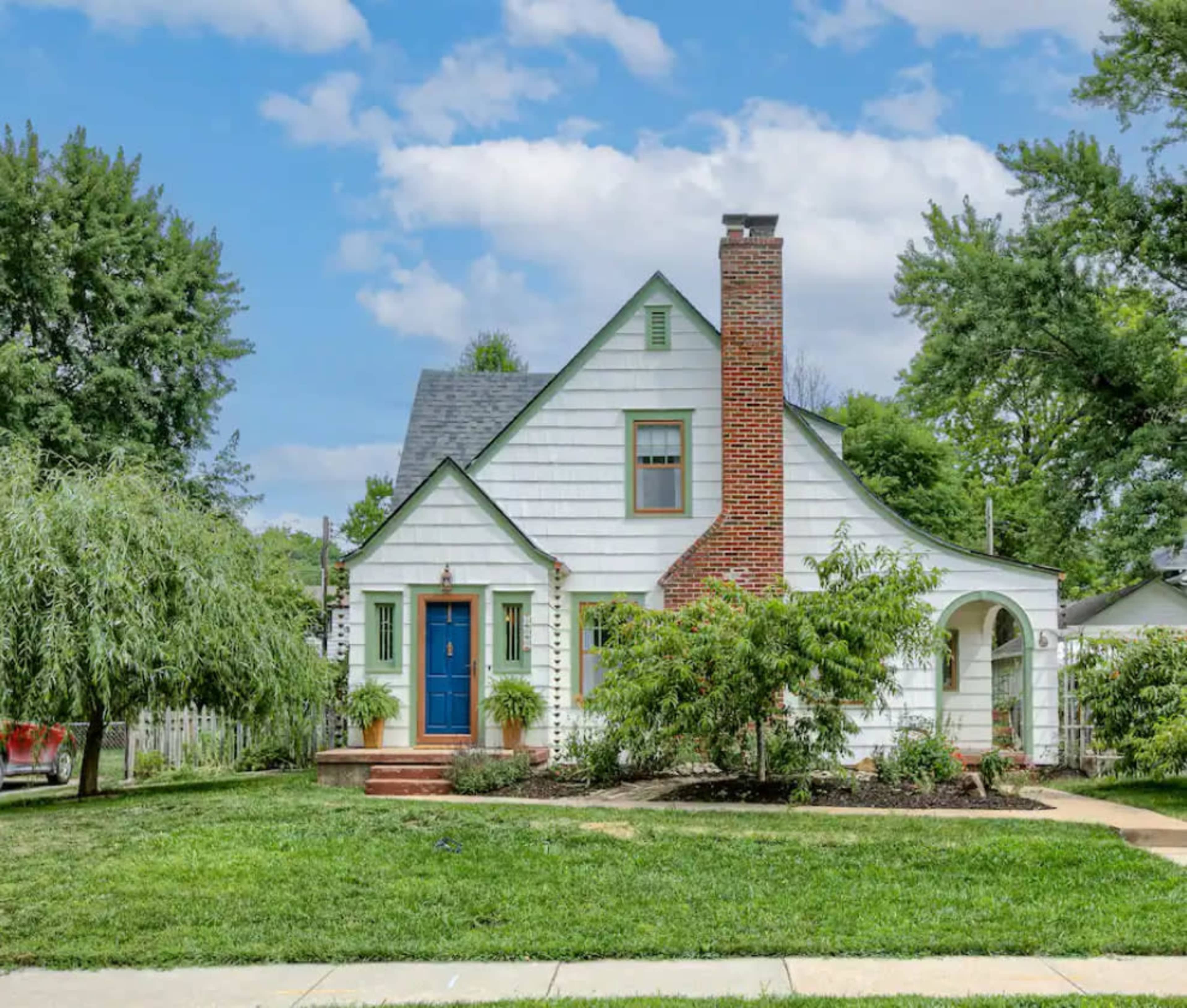 A two-story, white clapboard house features a blue front door, a brick chimney, and a well-maintained front yard with trees and shrubs.
