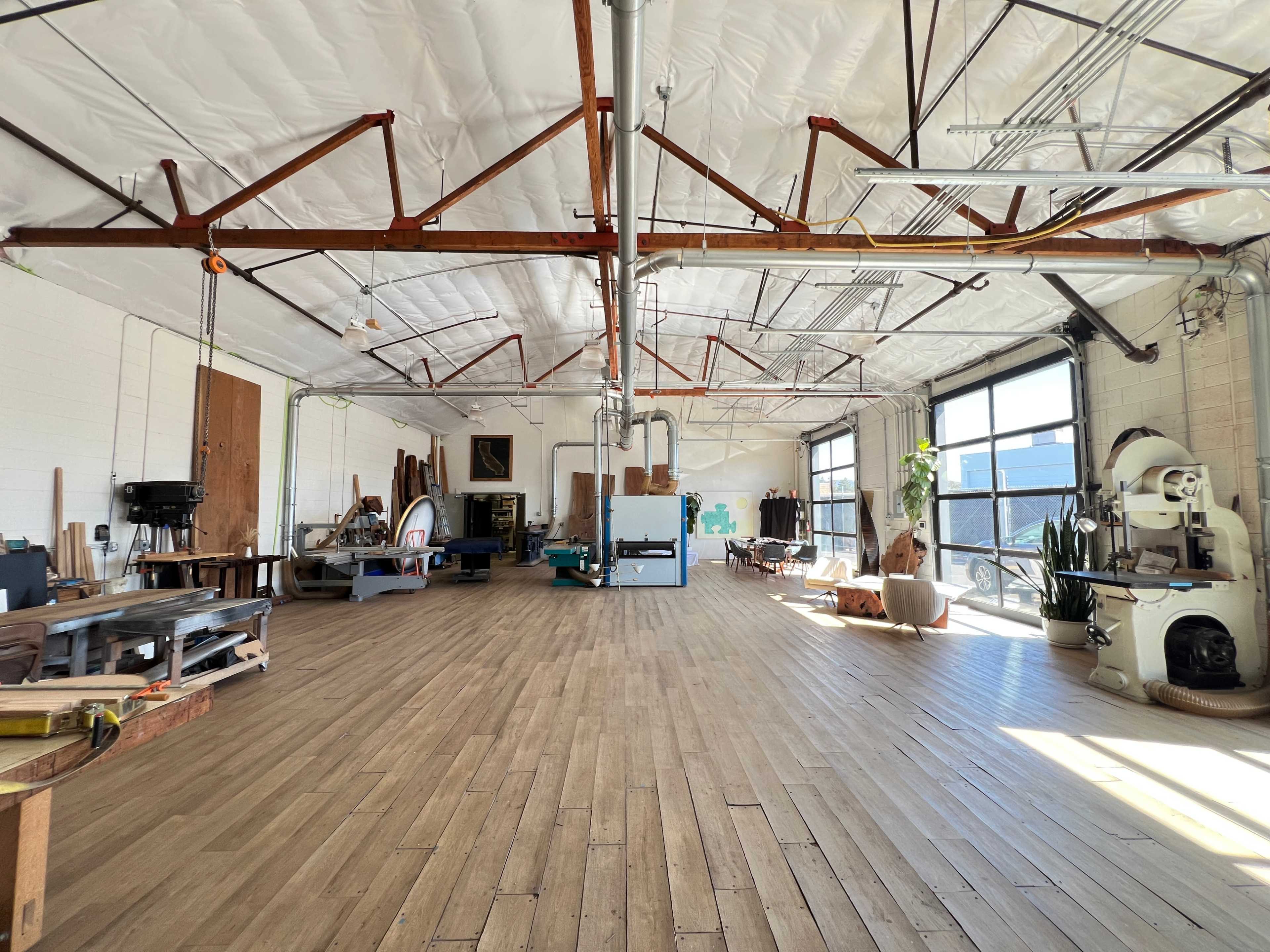 The image shows a spacious workshop with wooden floors, exposed beams, large windows, and various tools and furniture scattered throughout the area.