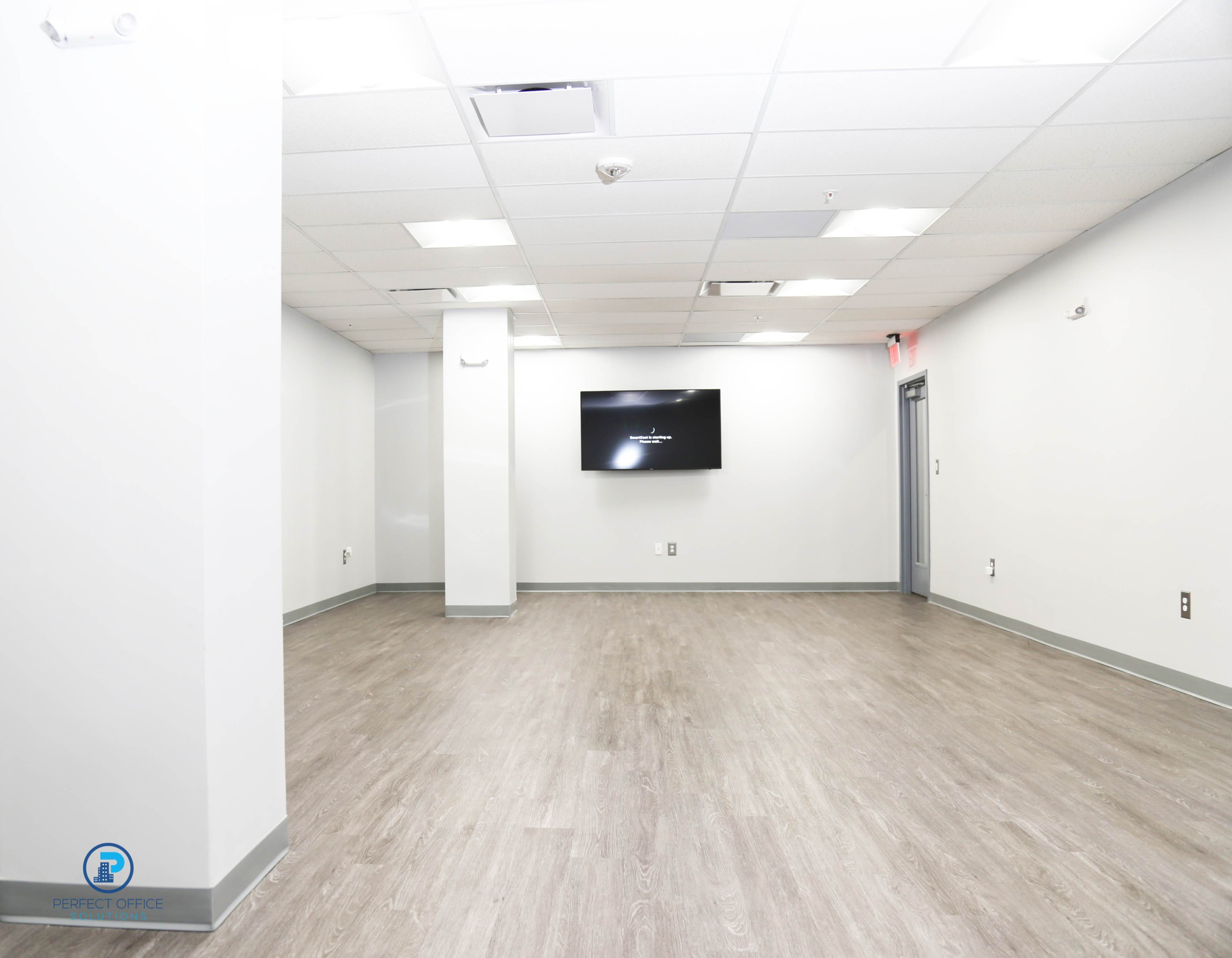 A spacious, empty office interior with light gray walls and a wooden floor, featuring a wall-mounted television.