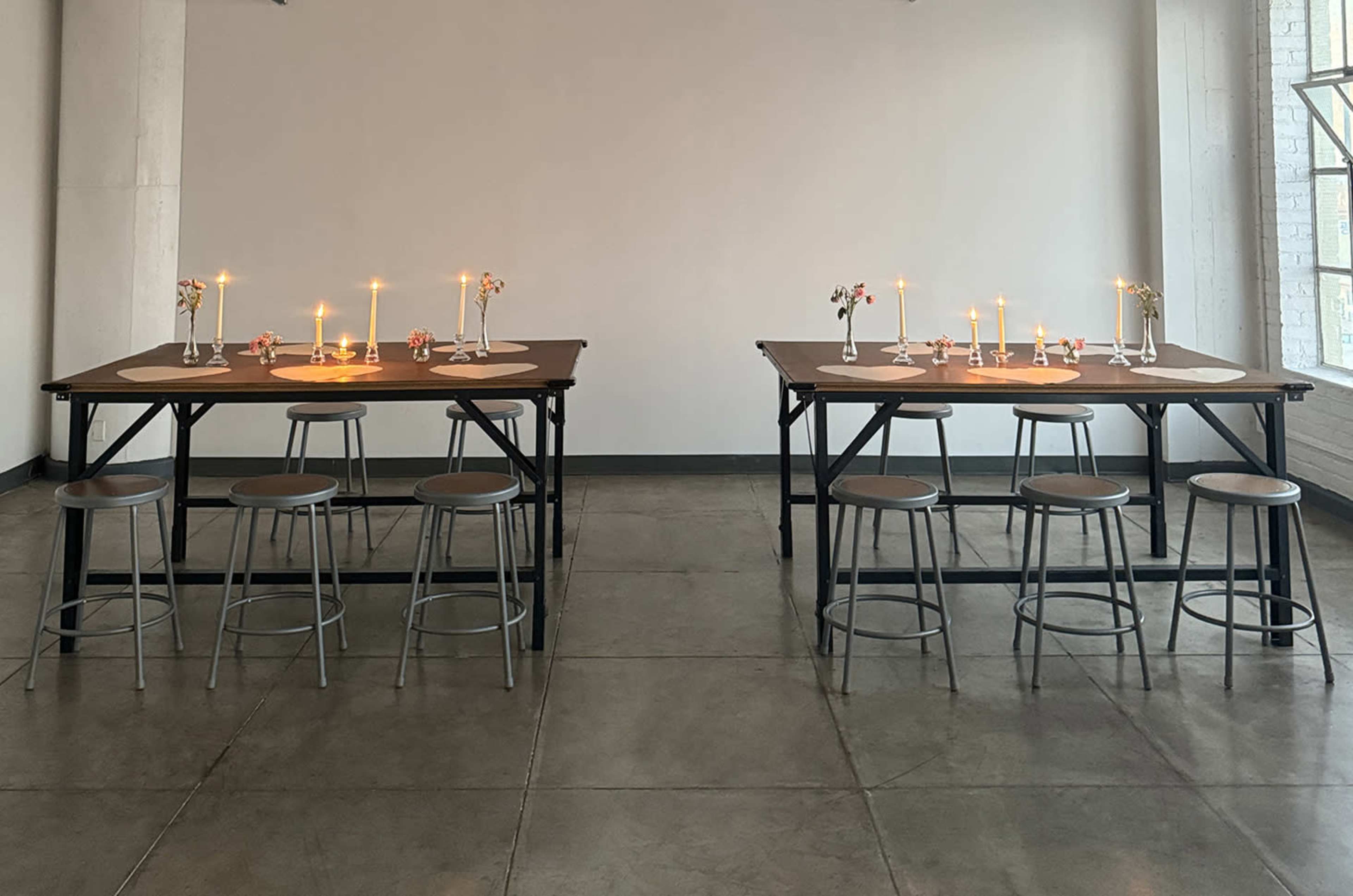 The image shows two tables arranged in a spacious room, each adorned with candles and small flower arrangements, surrounded by stools.