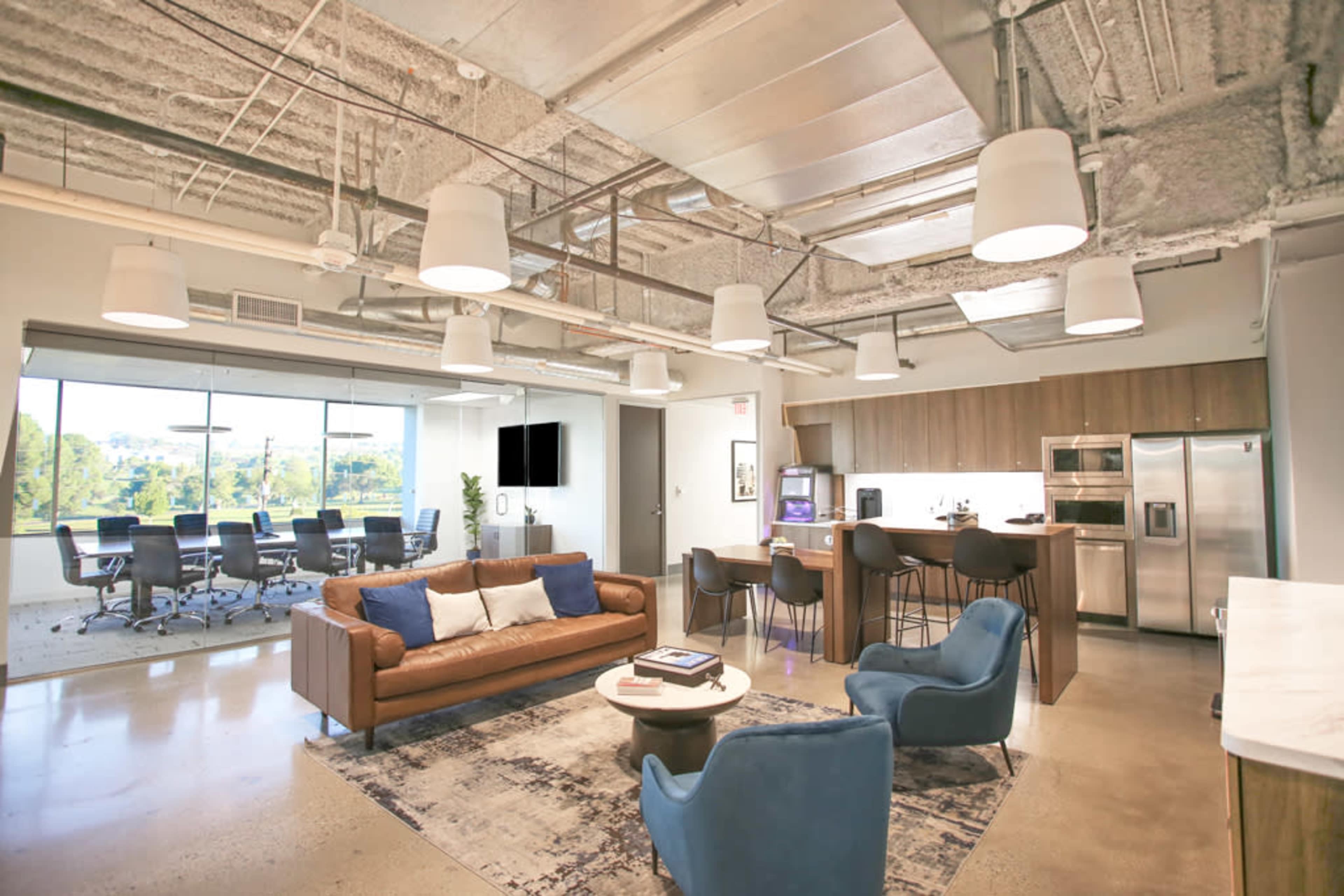 A modern office space featuring a lounge area with a sofa and chairs, an open kitchen, and a conference room with a large table visible through a glass wall.