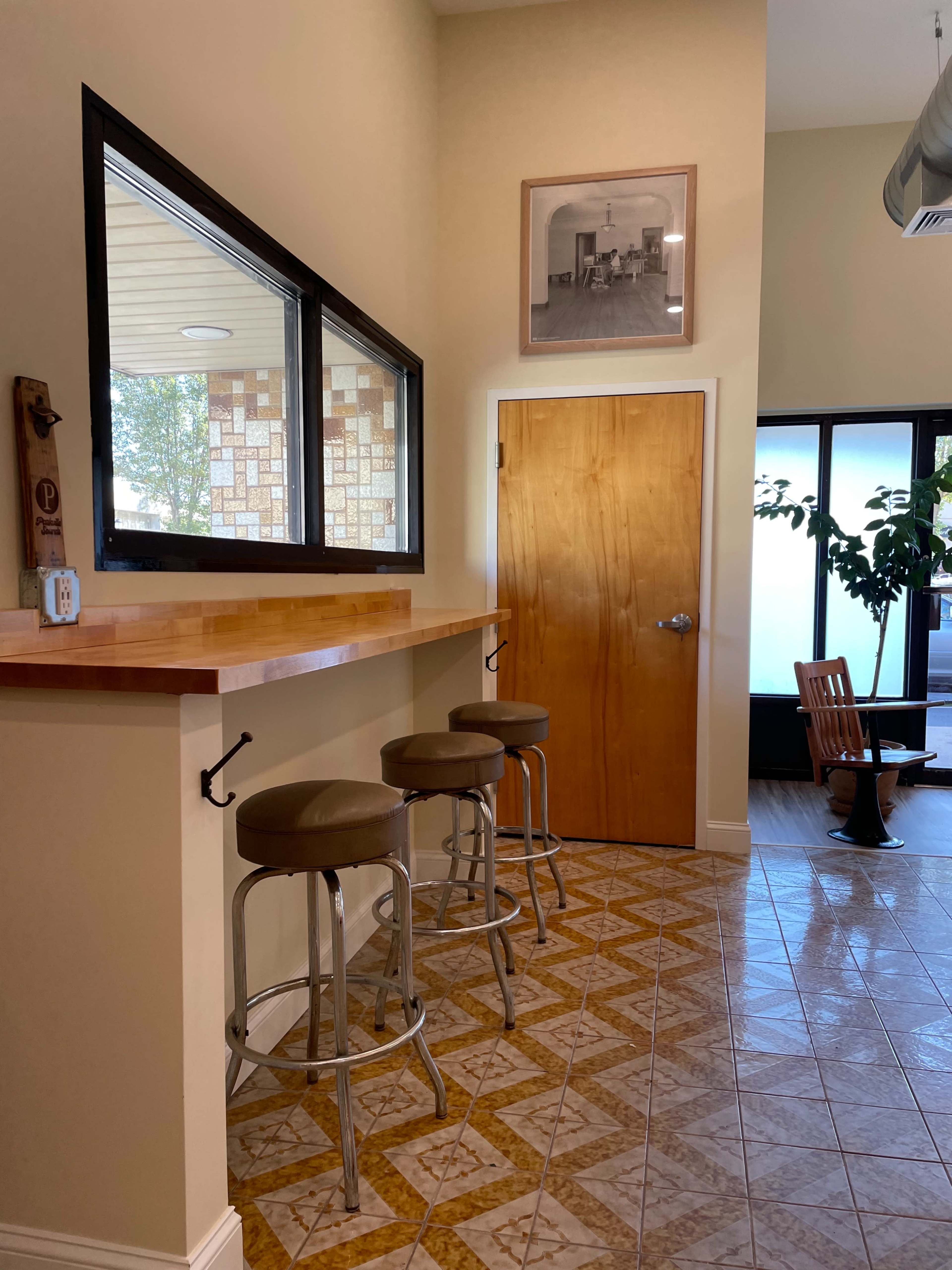 A bar area with three stools, a wooden countertop, and a door leading to another room, all set against a tiled floor and a framed photo on the wall.