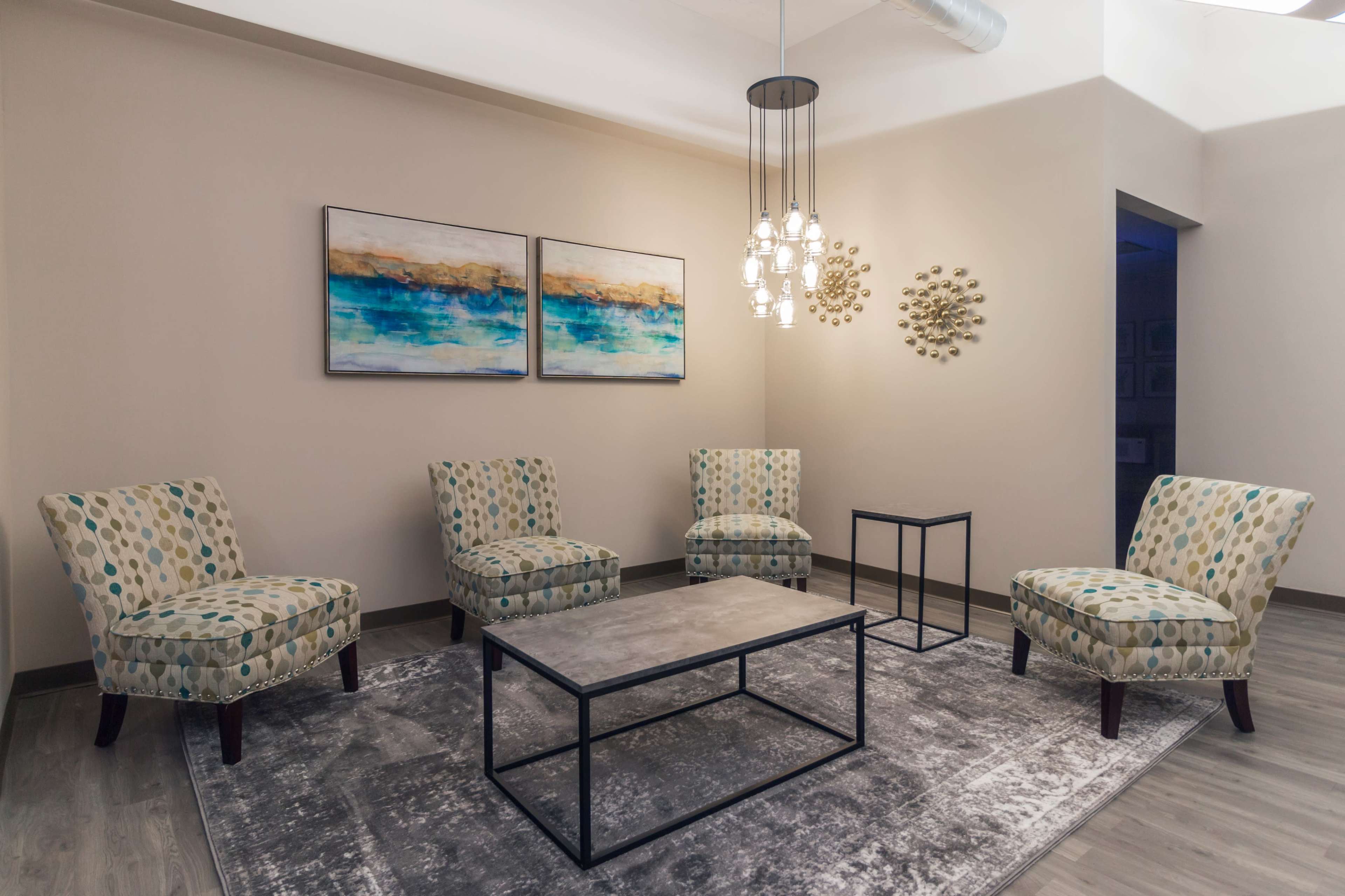 A modern waiting area features four patterned chairs arranged around a coffee table on a gray rug, with abstract artwork on the walls and a unique light fixture overhead.