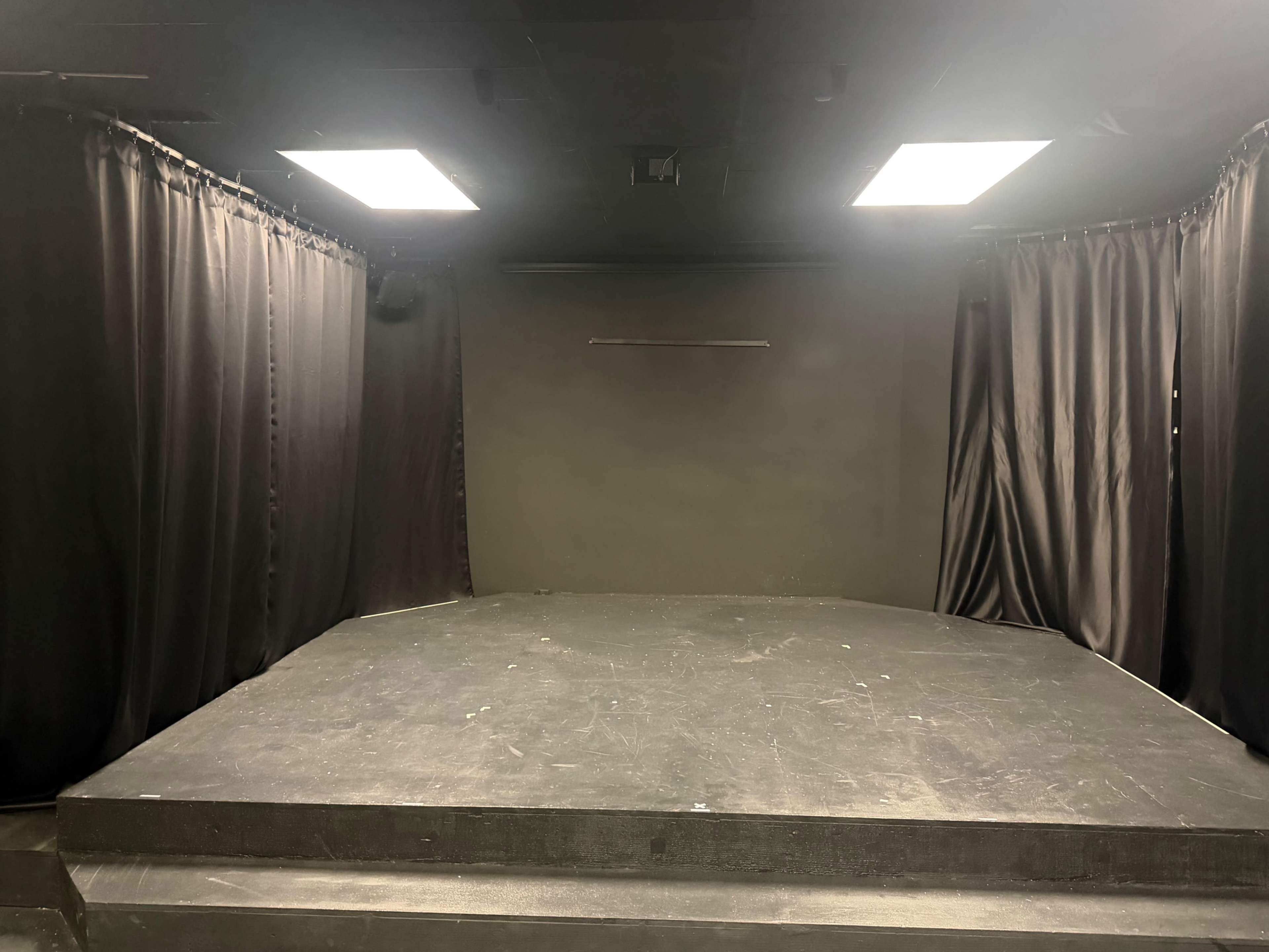 The image shows a small stage in a black curtain-draped room with spotlights overhead and a simple raised platform.