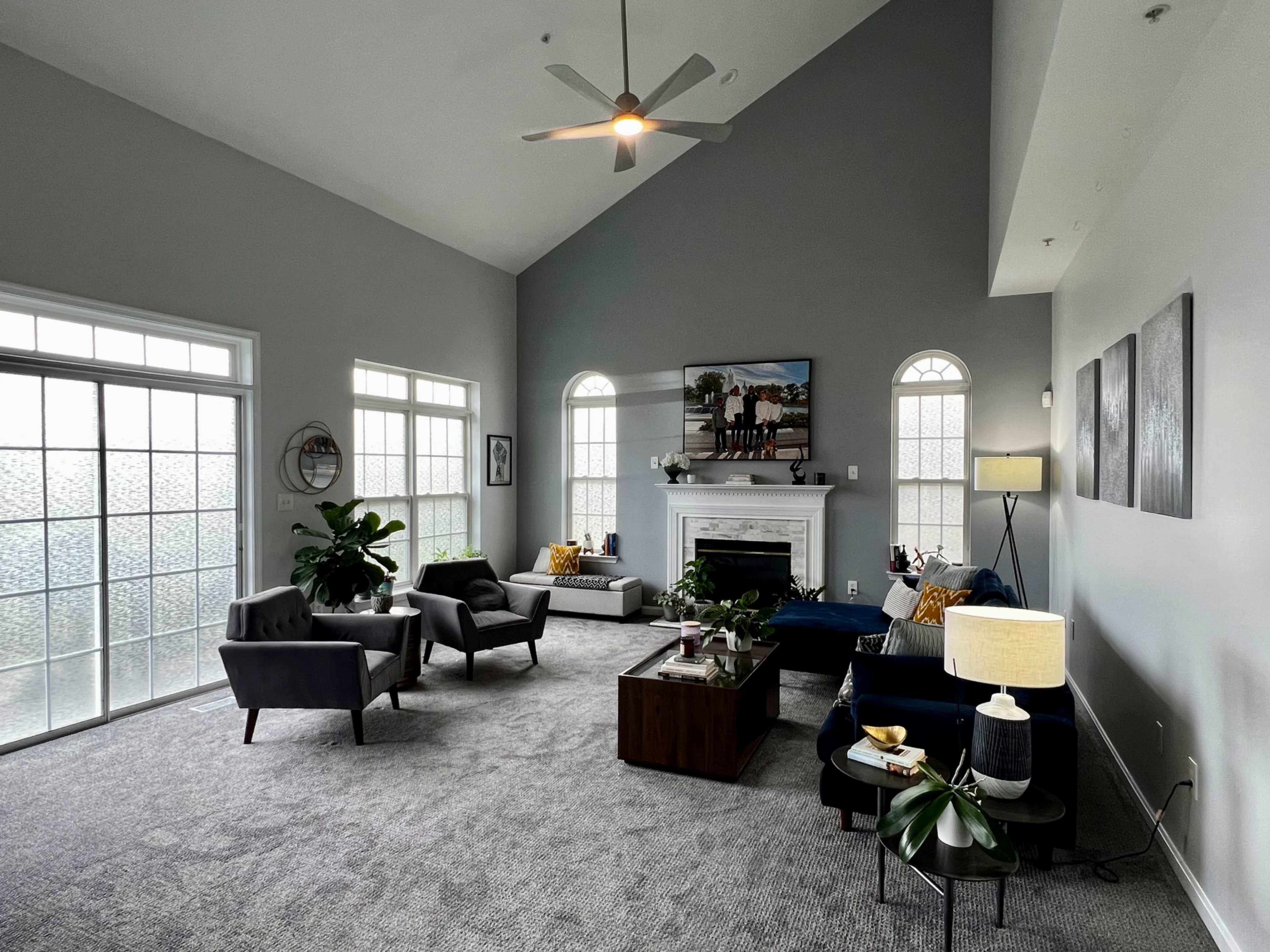 A spacious living room featuring a high ceiling, a fireplace, large windows, and modern furniture arranged around a coffee table.