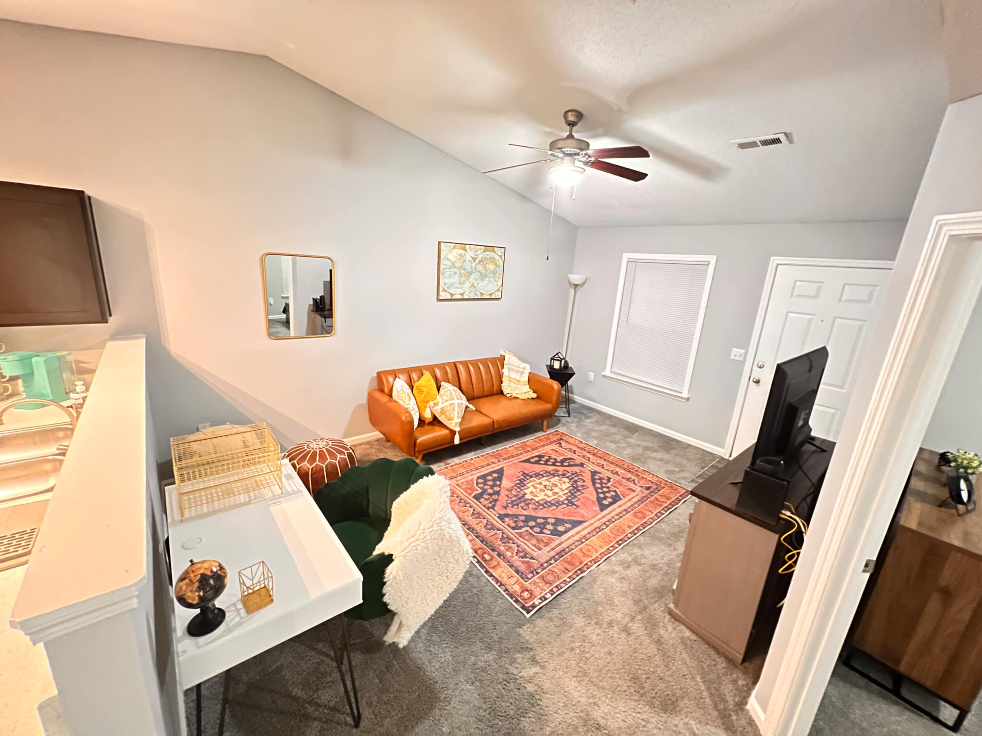 A cozy living room area with a brown sofa, a patterned rug, and a small dining table, all illuminated by natural light from a nearby window.