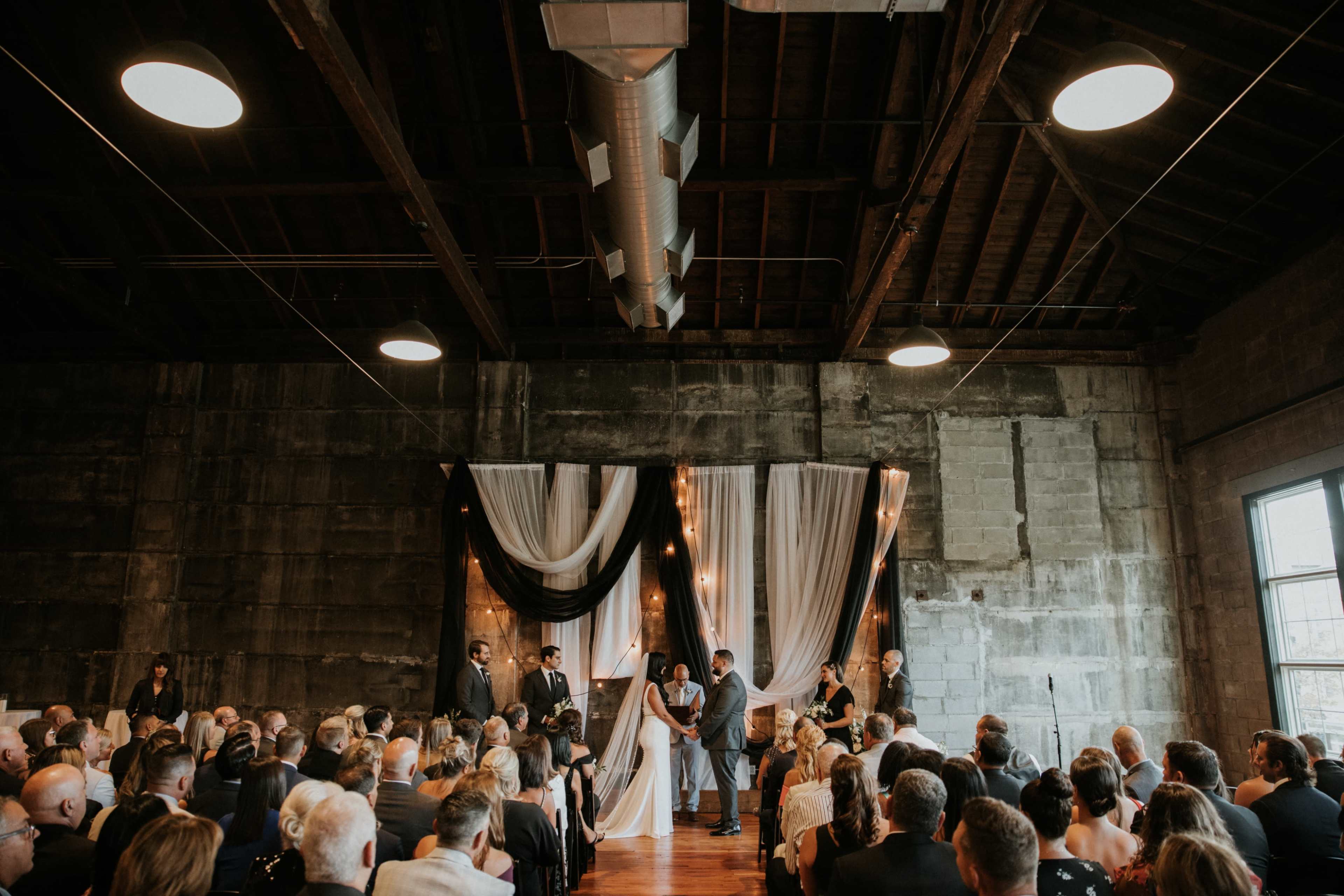 A wedding ceremony takes place indoors with guests seated in rows, facing a decorated altar featuring draped fabric and lighting.