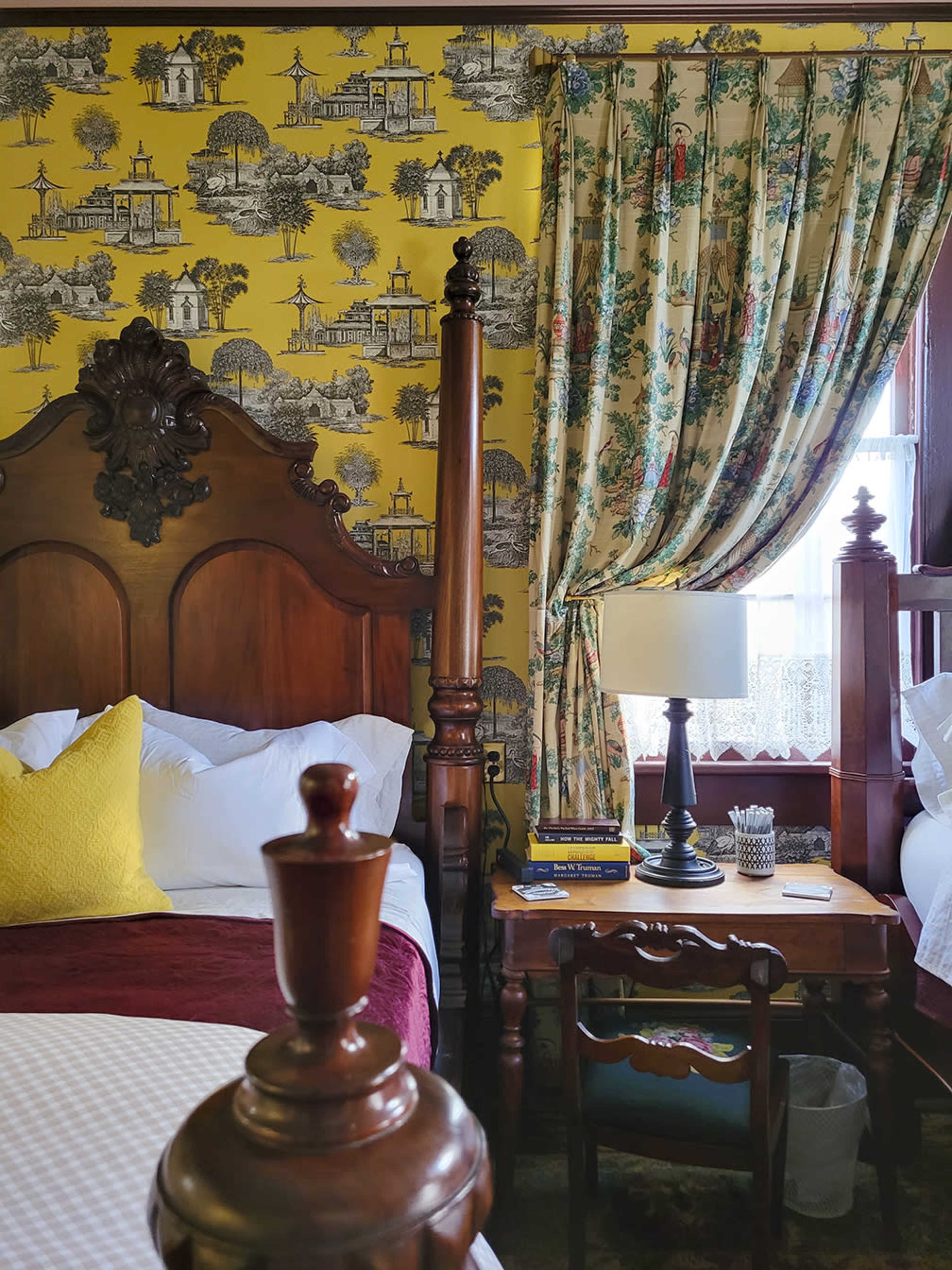 A cozy bedroom featuring a yellow wallpaper with a countryside motif, a wooden bed with a decorative headboard, a side table with a lamp, and floral curtains.