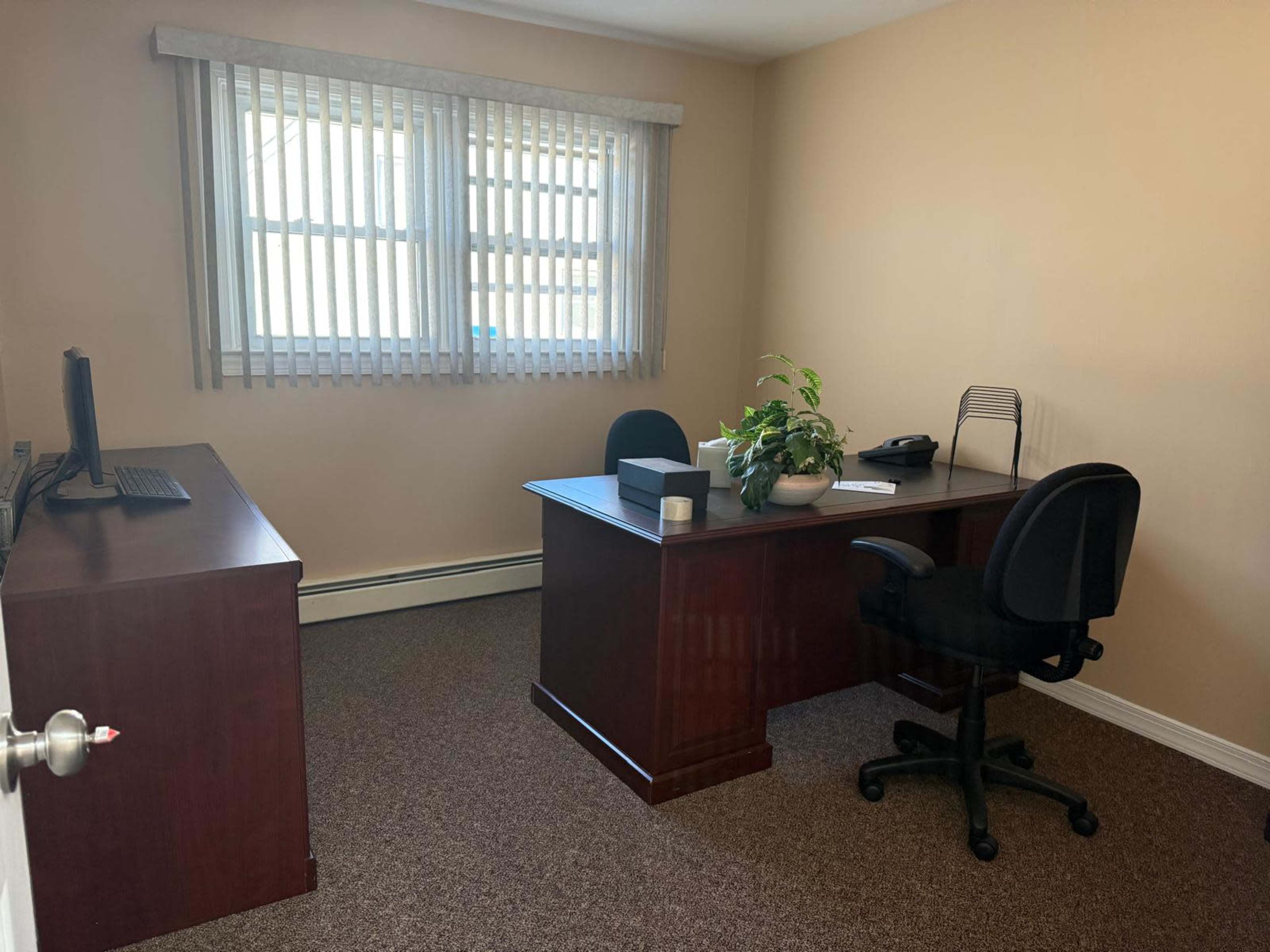 The image shows a simple office space with a wooden desk, a computer, a chair, and a potted plant, illuminated by natural light from two windows.