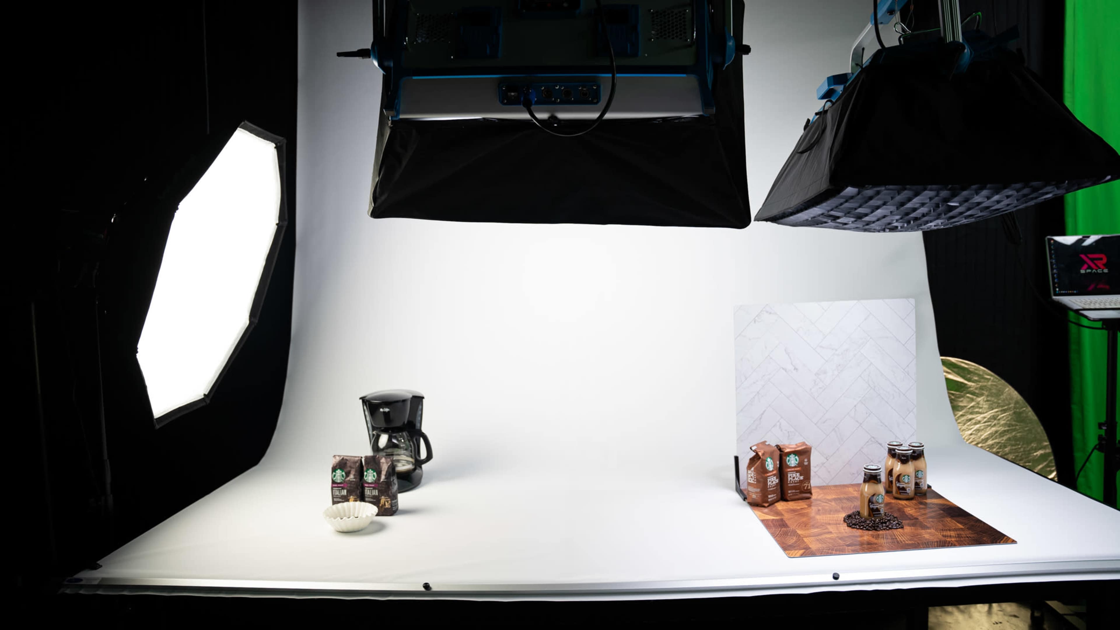 The image shows a photography studio setup with a coffee maker, coffee boxes, and cans arranged on a wooden surface against a white backdrop.