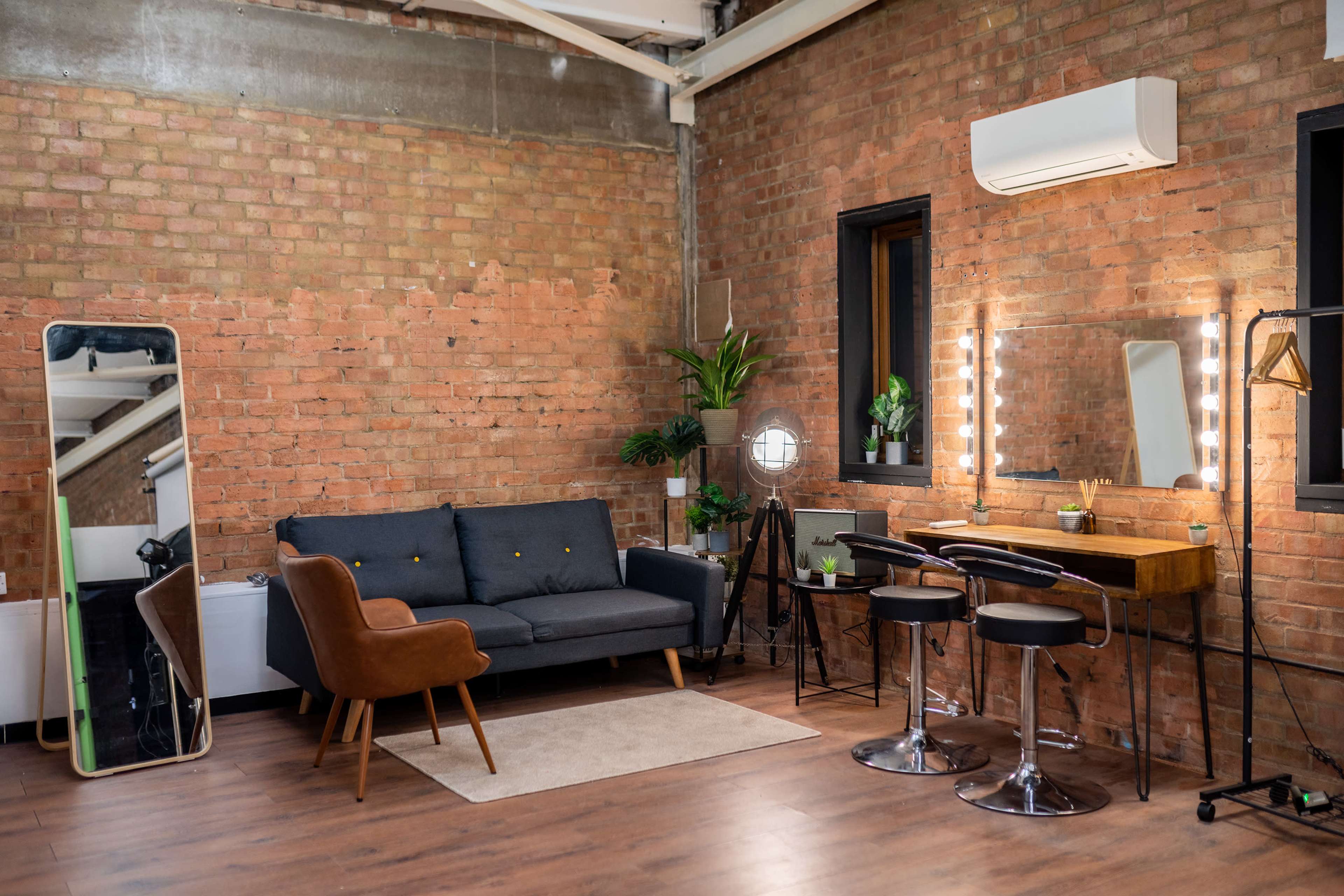 A modern interior featuring a gray couch, a large mirror, a wooden table with a lamp, and two bar stools in a room with exposed brick walls.
