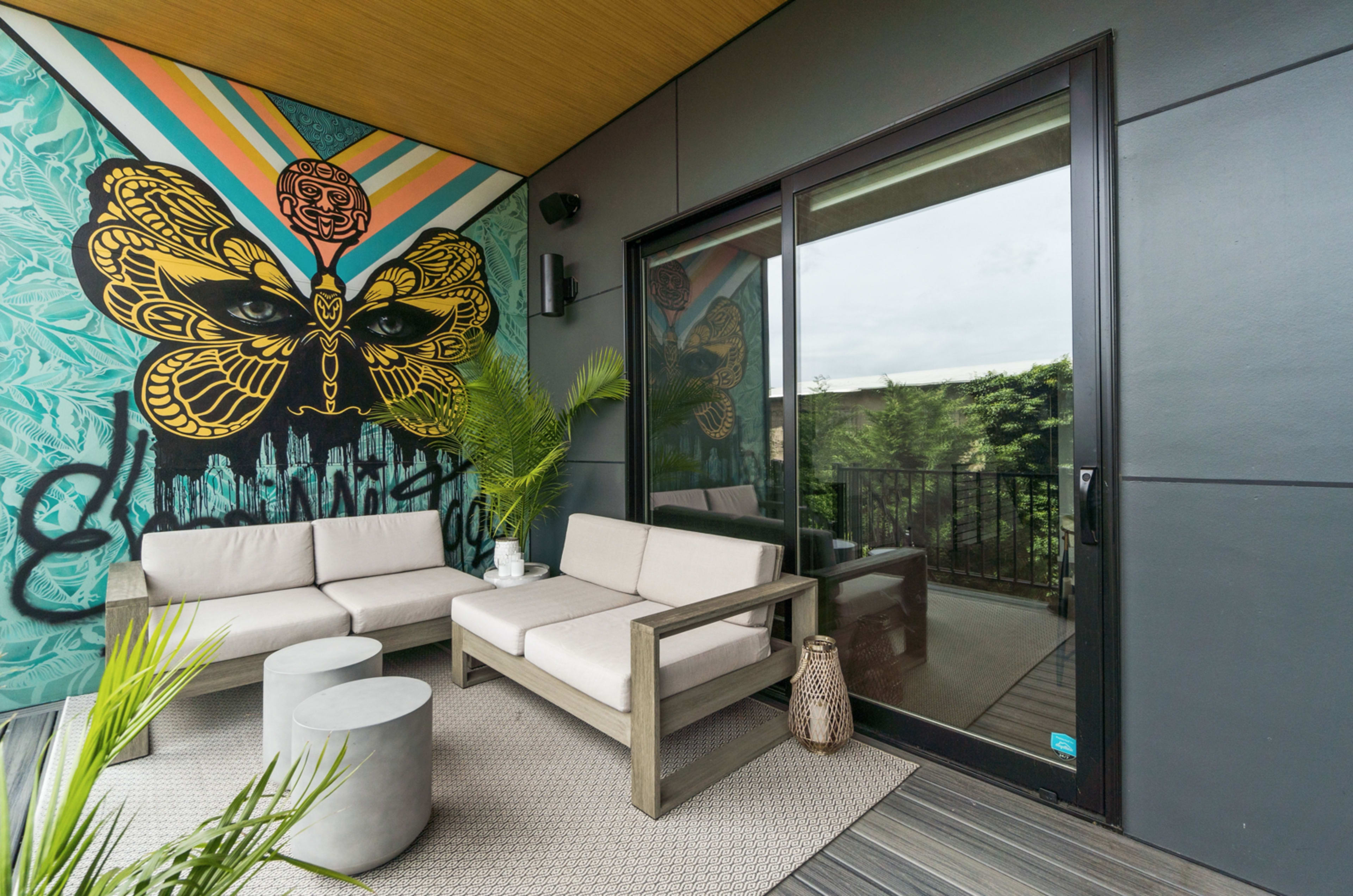 A modern balcony features a colorful butterfly mural, two light-colored sofas, a round coffee table, and greenery accompanying the glass sliding door.