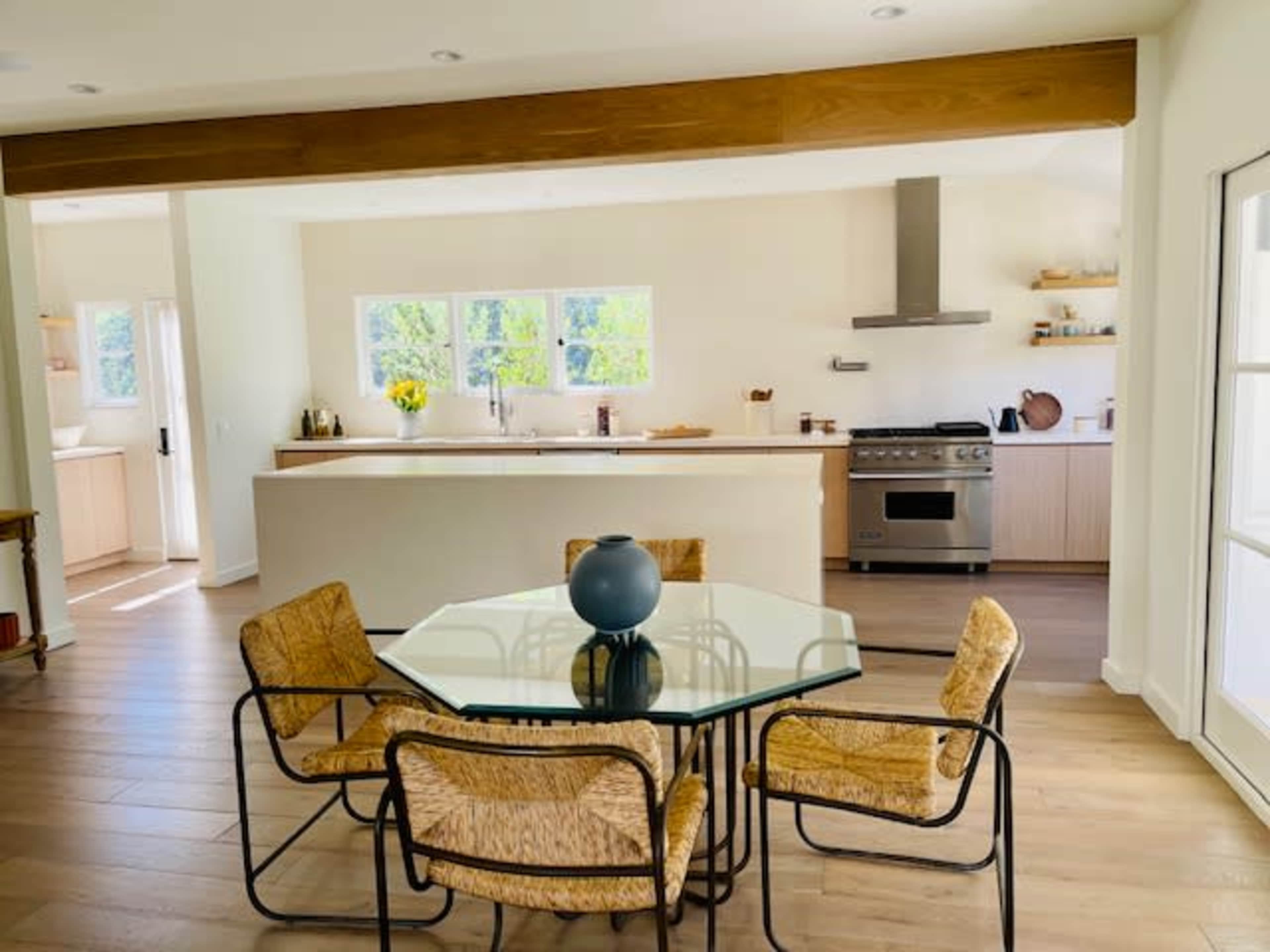 A dining area features a glass-top table surrounded by four woven chairs, adjacent to a modern kitchen with stainless steel appliances and natural light from large windows.