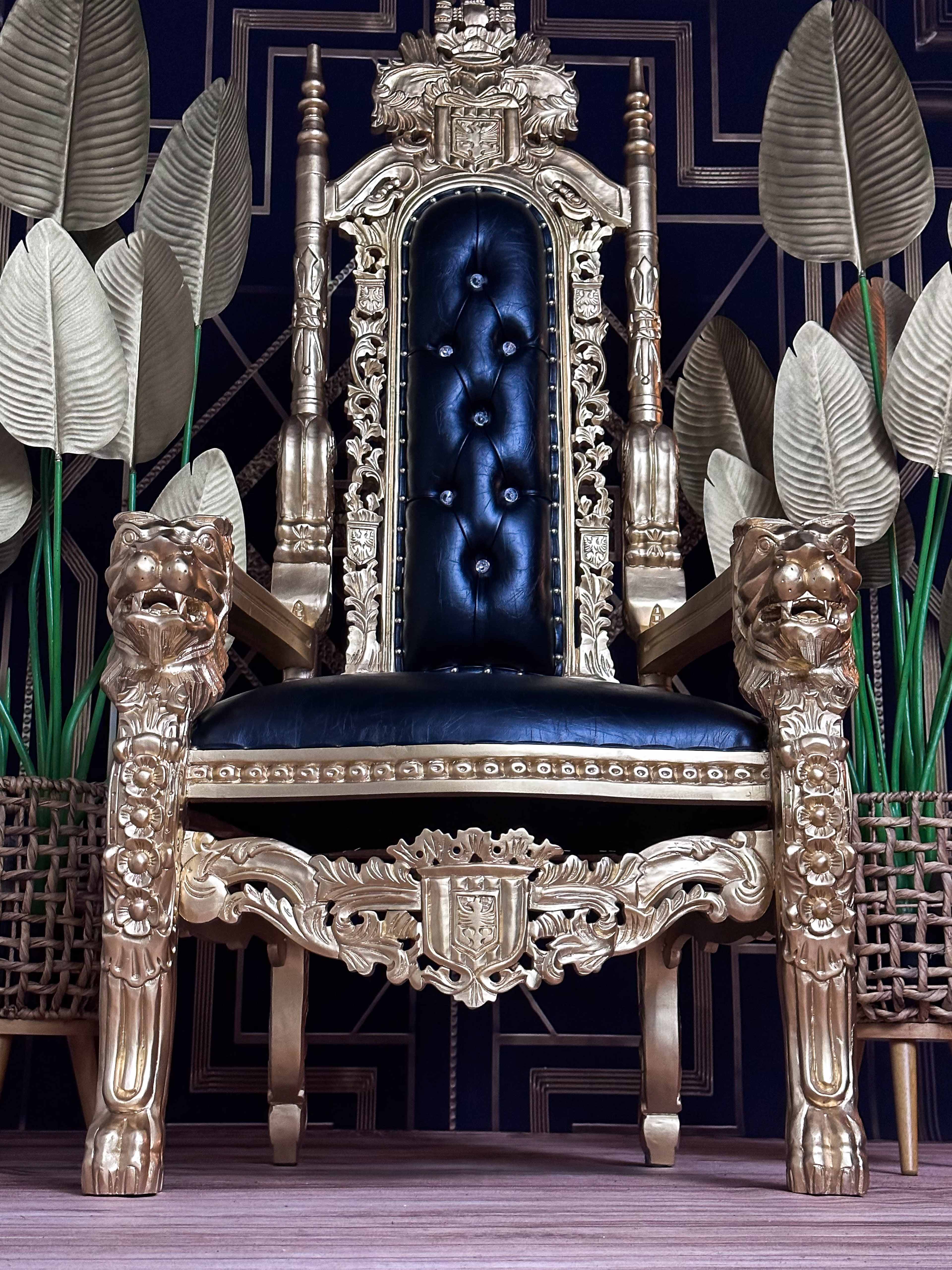 A large, ornate golden throne with a black cushioned seat, flanked by decorative leaves in the background.