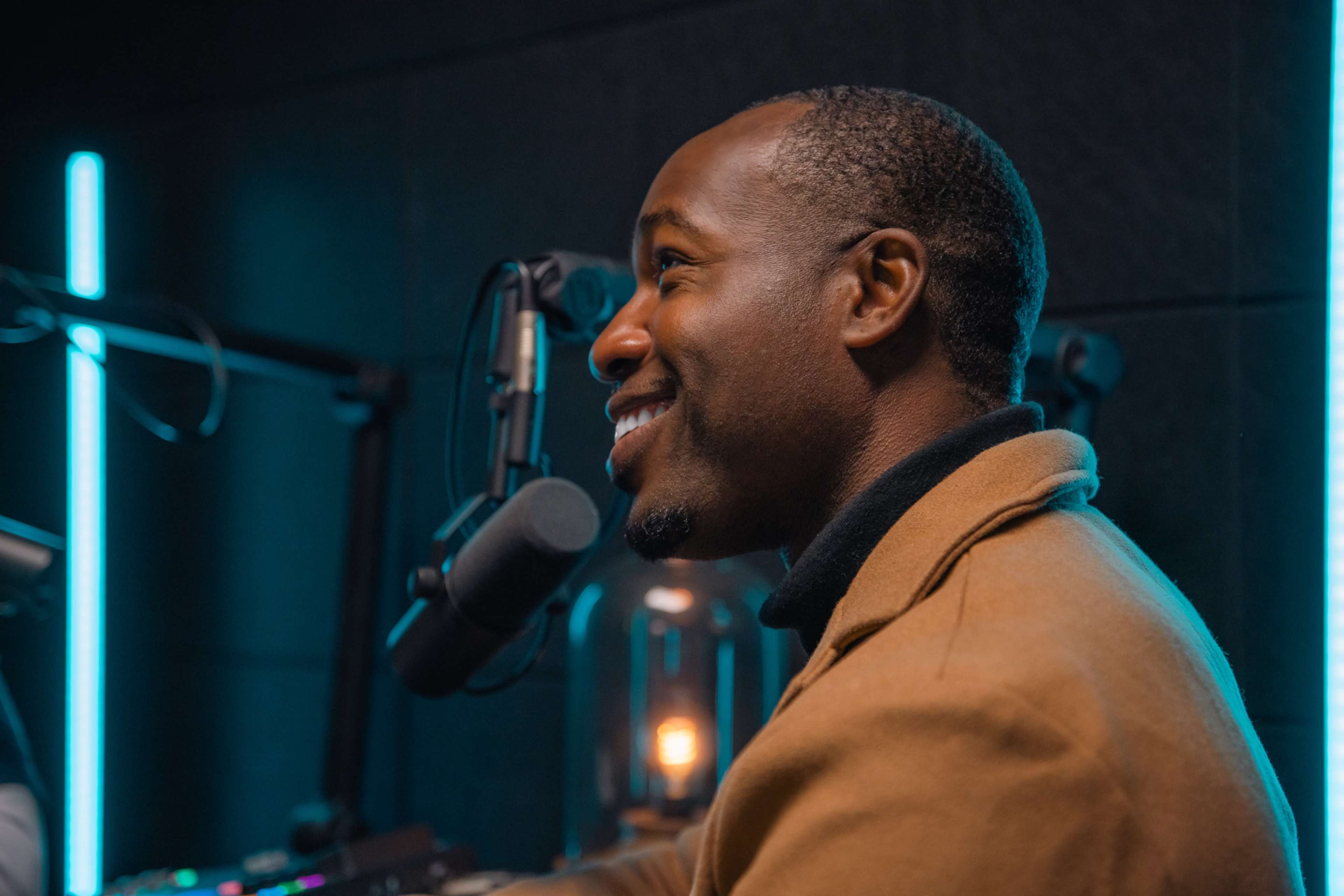 A man is sitting in a recording studio, smiling while speaking into a microphone, with blue neon lights illuminating the background.