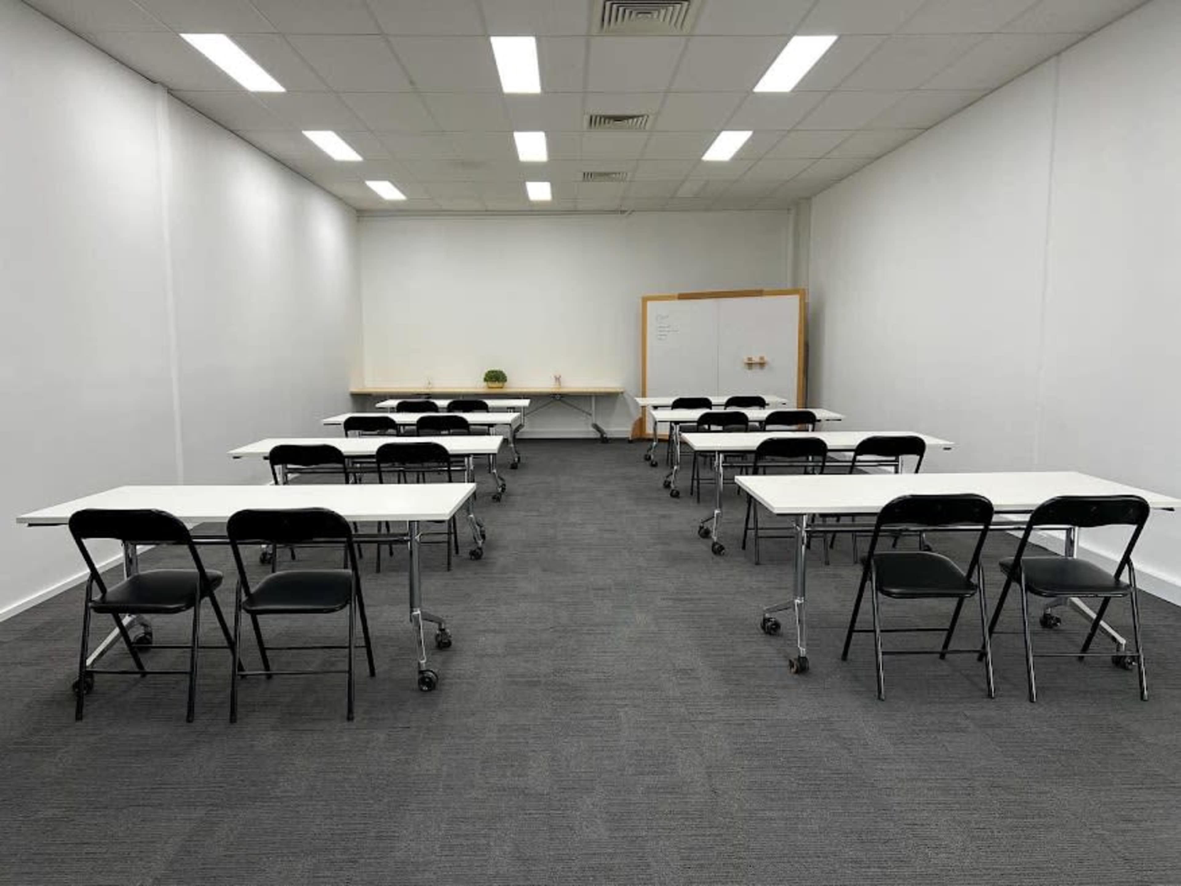 A brightly lit classroom with several rows of movable tables and chairs arranged in a neat layout against white walls.