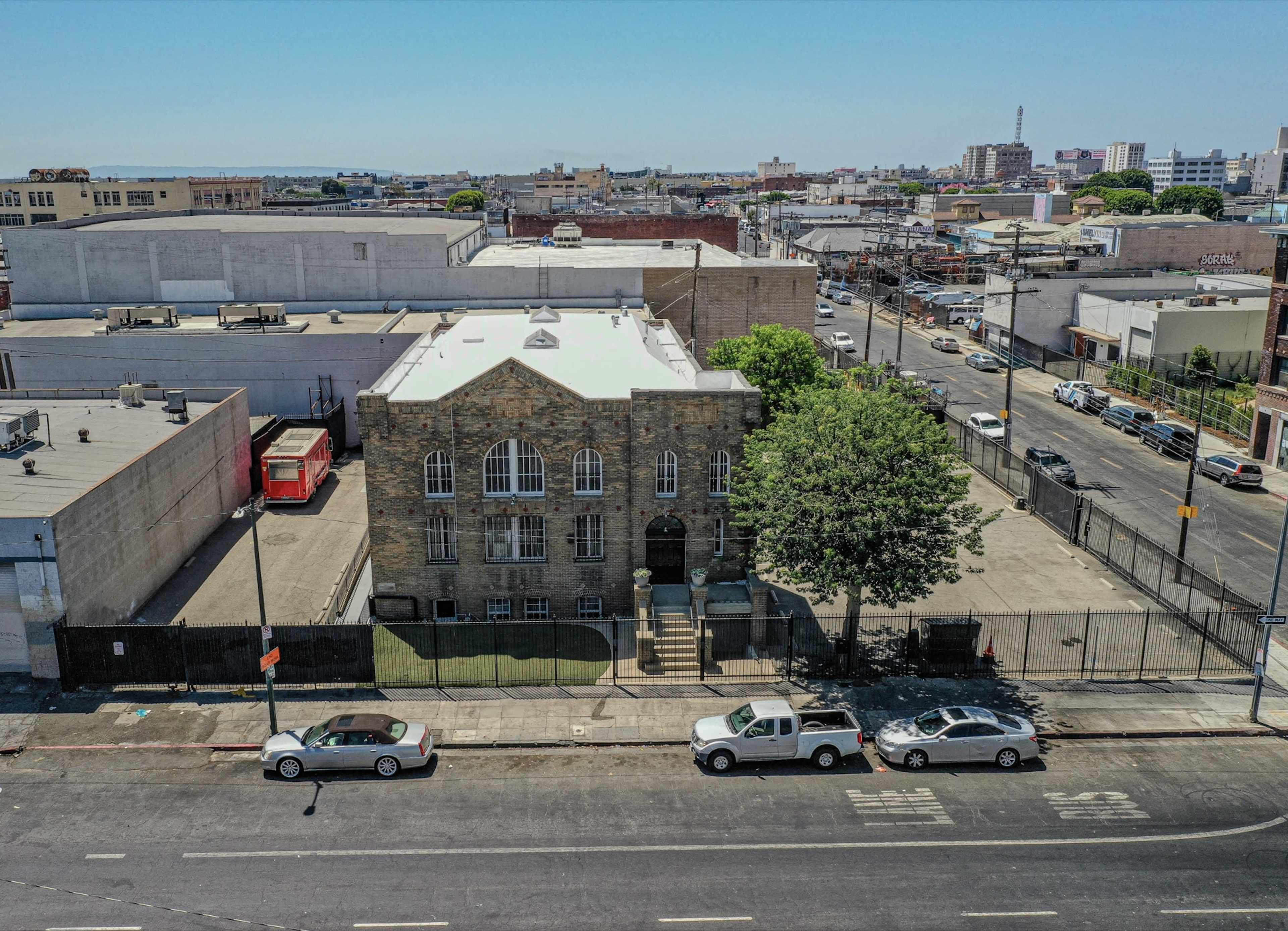 The image shows a large stone building with arched windows, surrounded by a fenced area and parked cars on a city street.