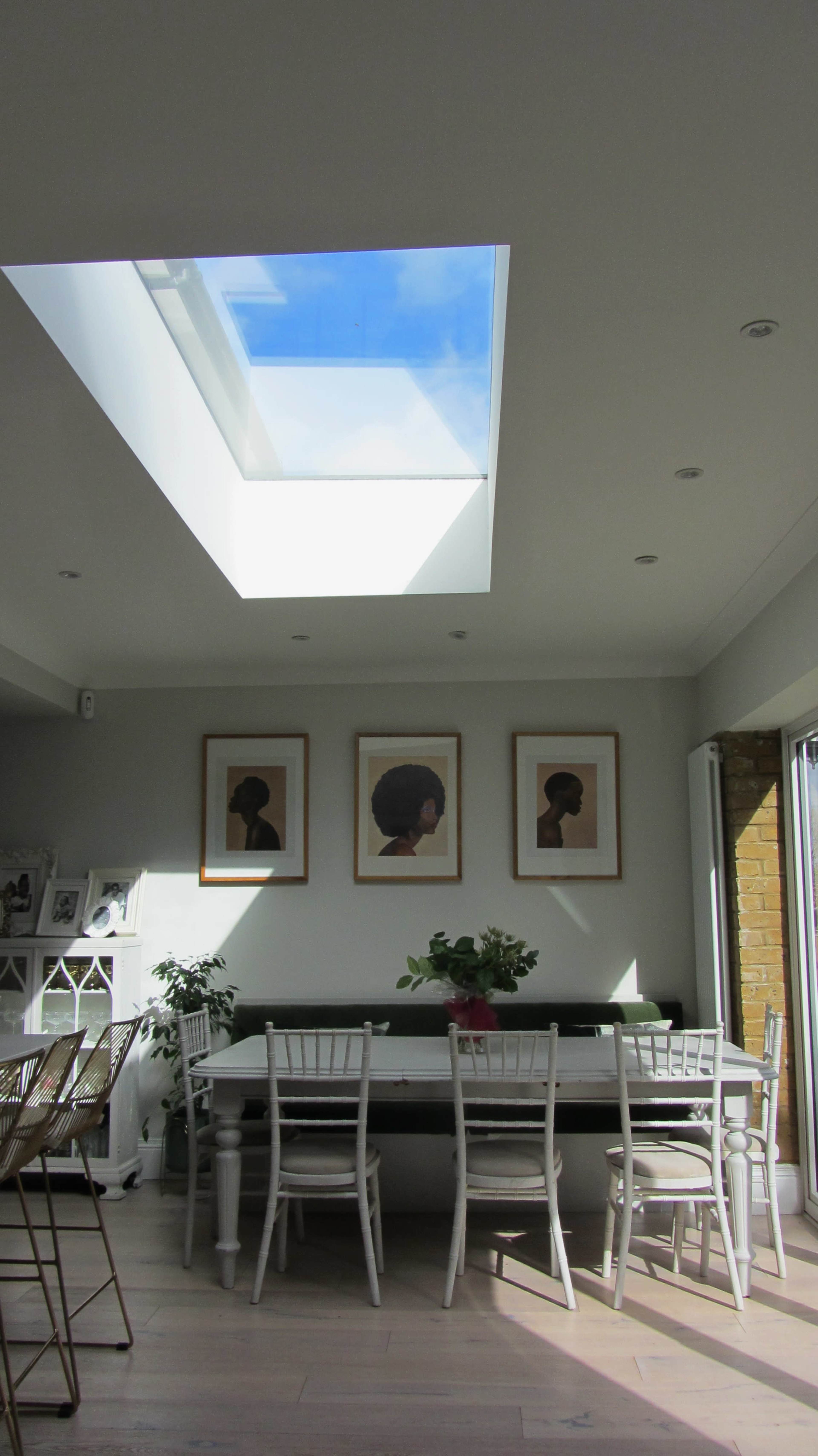 The image shows a bright room with a skylight, a long table surrounded by white chairs, and three framed artworks of profile silhouettes on the wall.