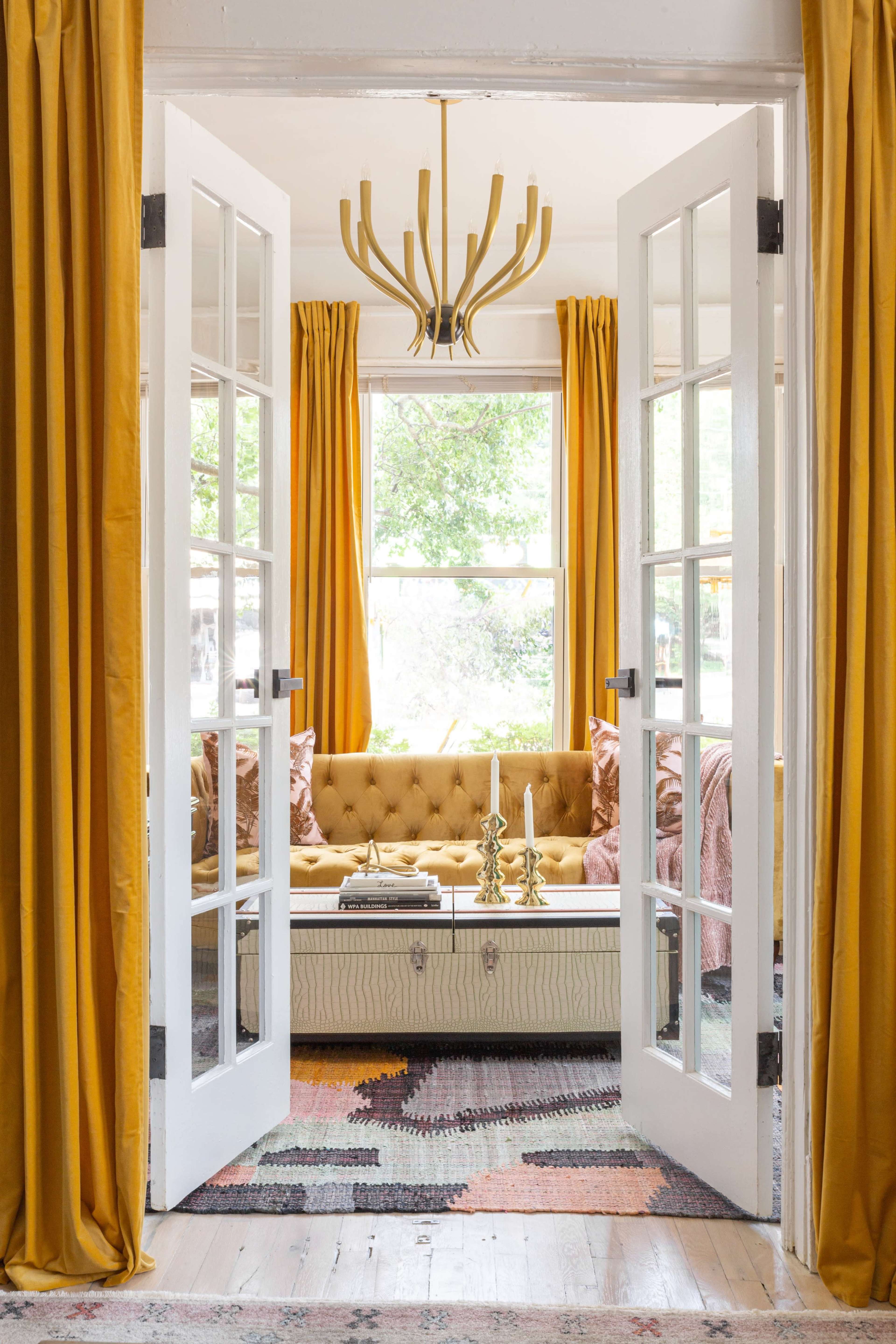 A living room featuring a tufted couch, yellow curtains, and a chandelier is visible through open French doors.