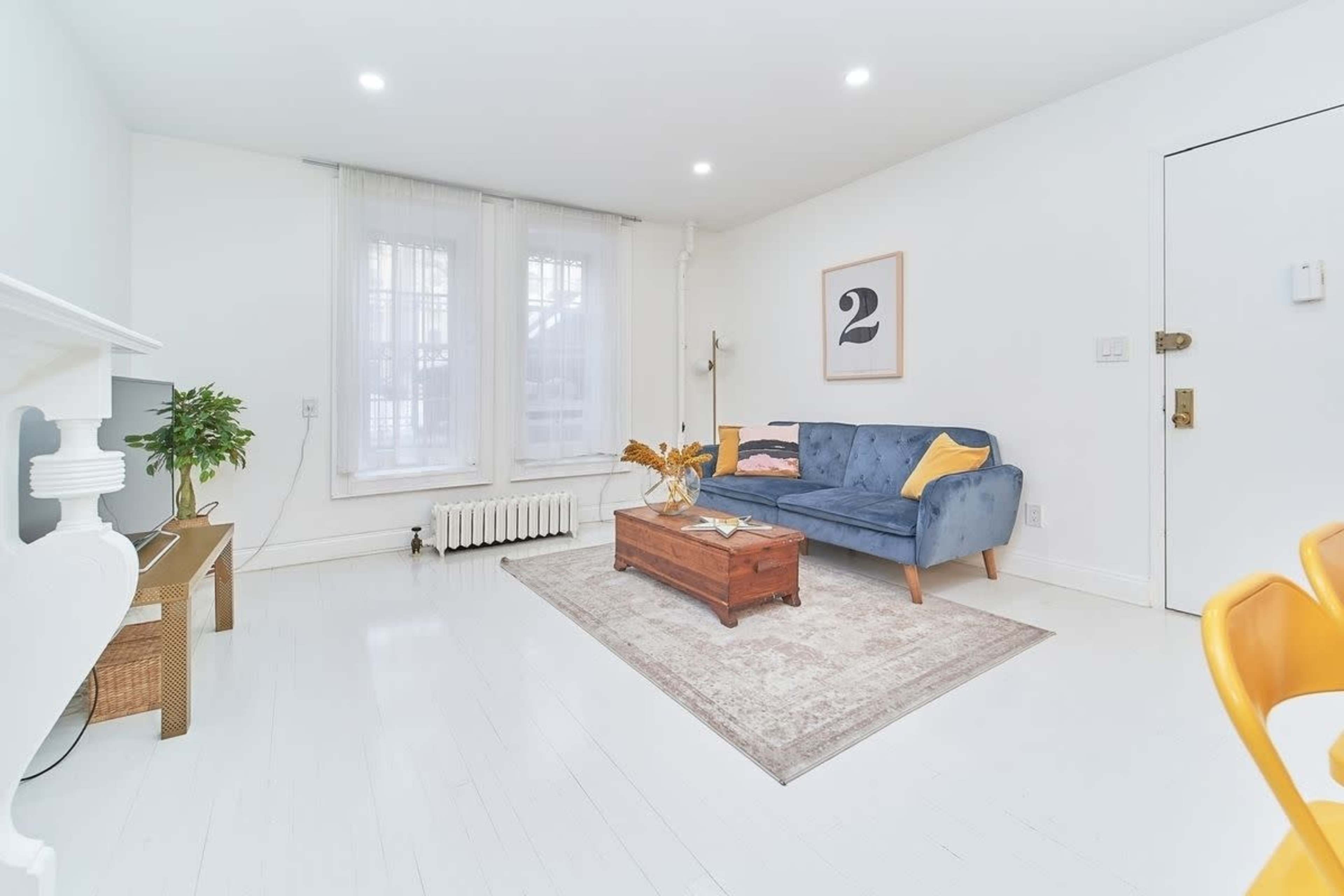 The image shows a modern living room with a blue sofa, a wooden coffee table, a large rug, and bright white walls, featuring large windows that allow natural light.